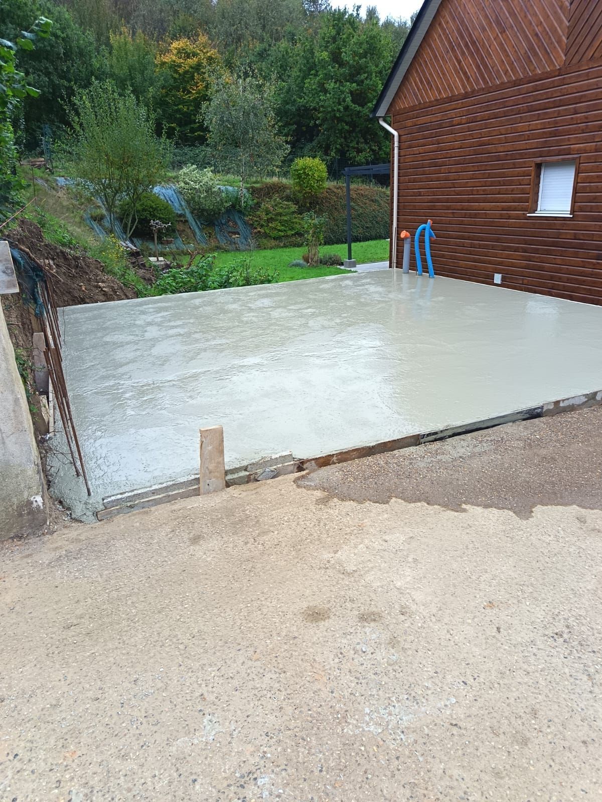 Dalle de béton fraîchement coulée à côté d'une maison en briques et d'une allée de gravier, avec un jardin boisé derrière.