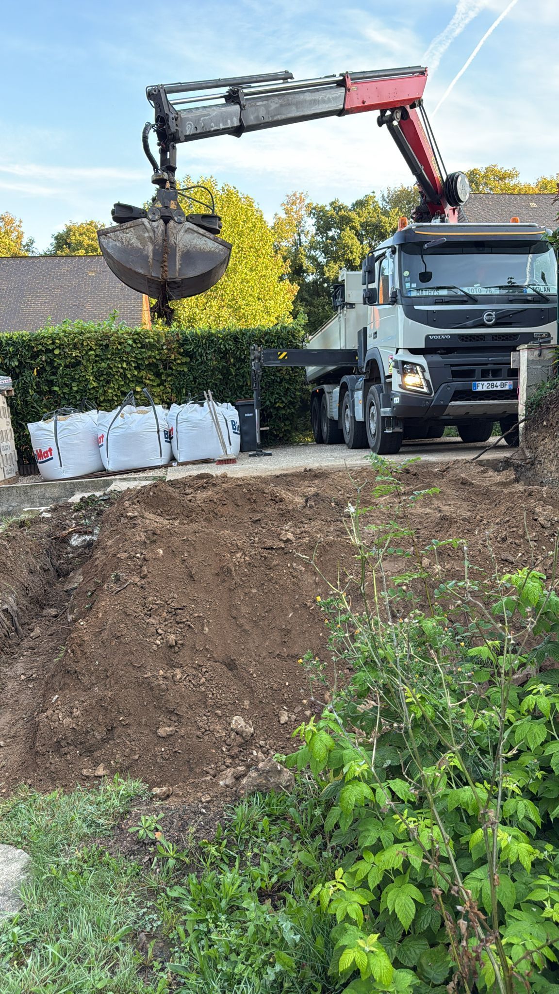 Grue montée sur camion soulevant un grand godet à côté d'un talus de terre, avec des barrières blanches et des arbres en arrière-plan.