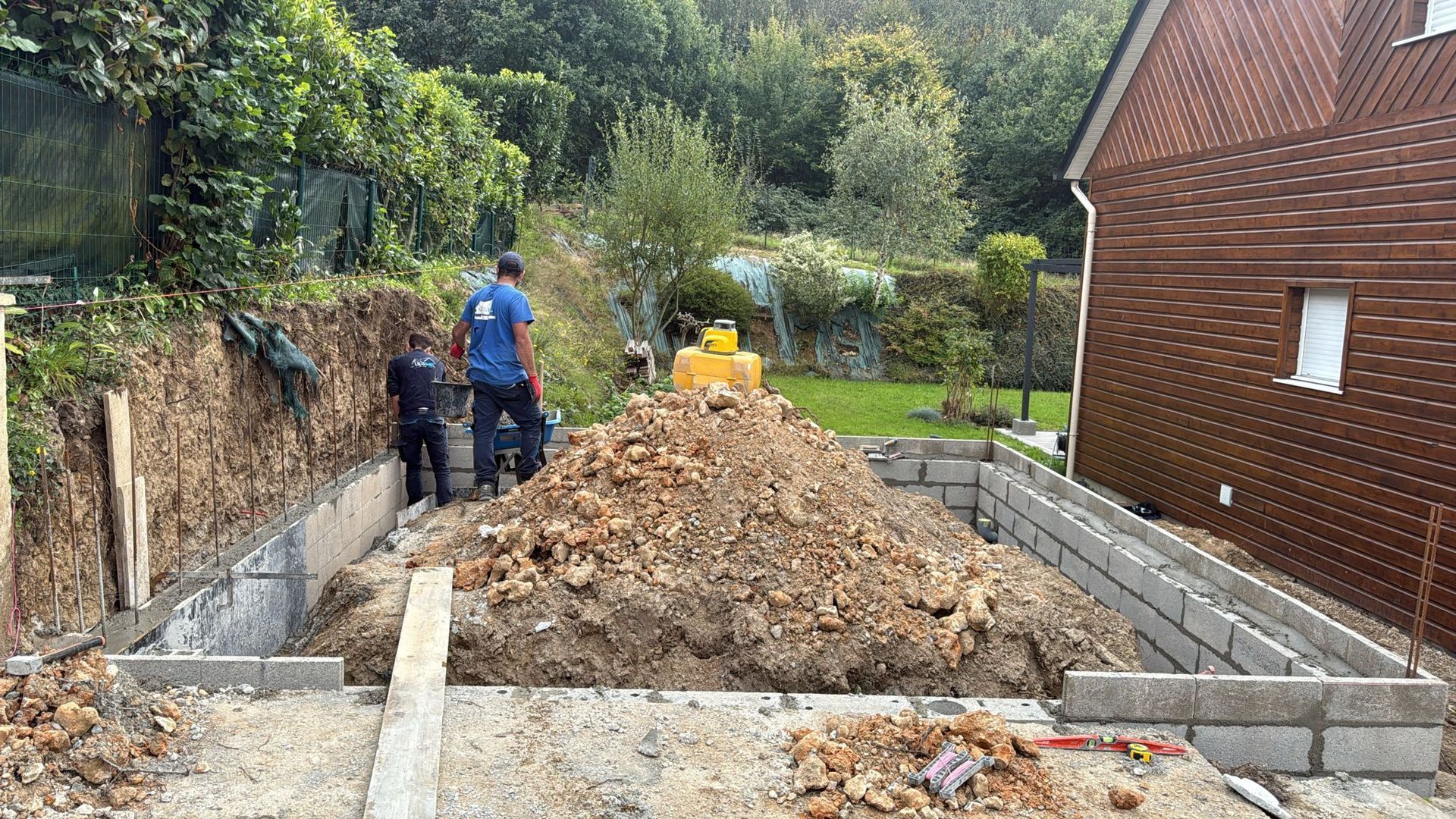 Des ouvriers creusent une tranchée sur un chantier de construction, à côté d'une maison en briques, avec un gros tas de terre au centre.