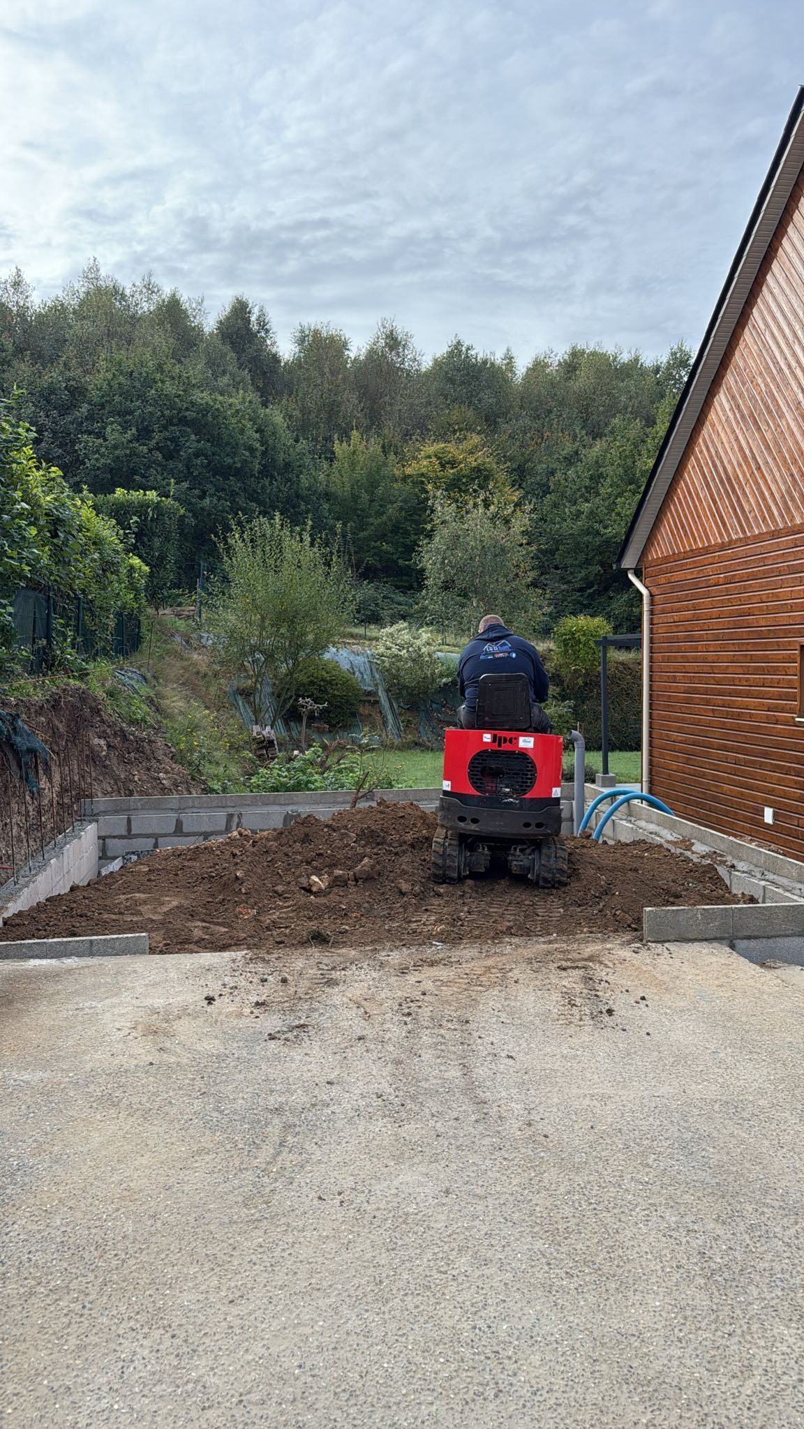 Petite machine de chantier sur du gravier dans un jardin, avec un versant boisé et un mur de briques à proximité.