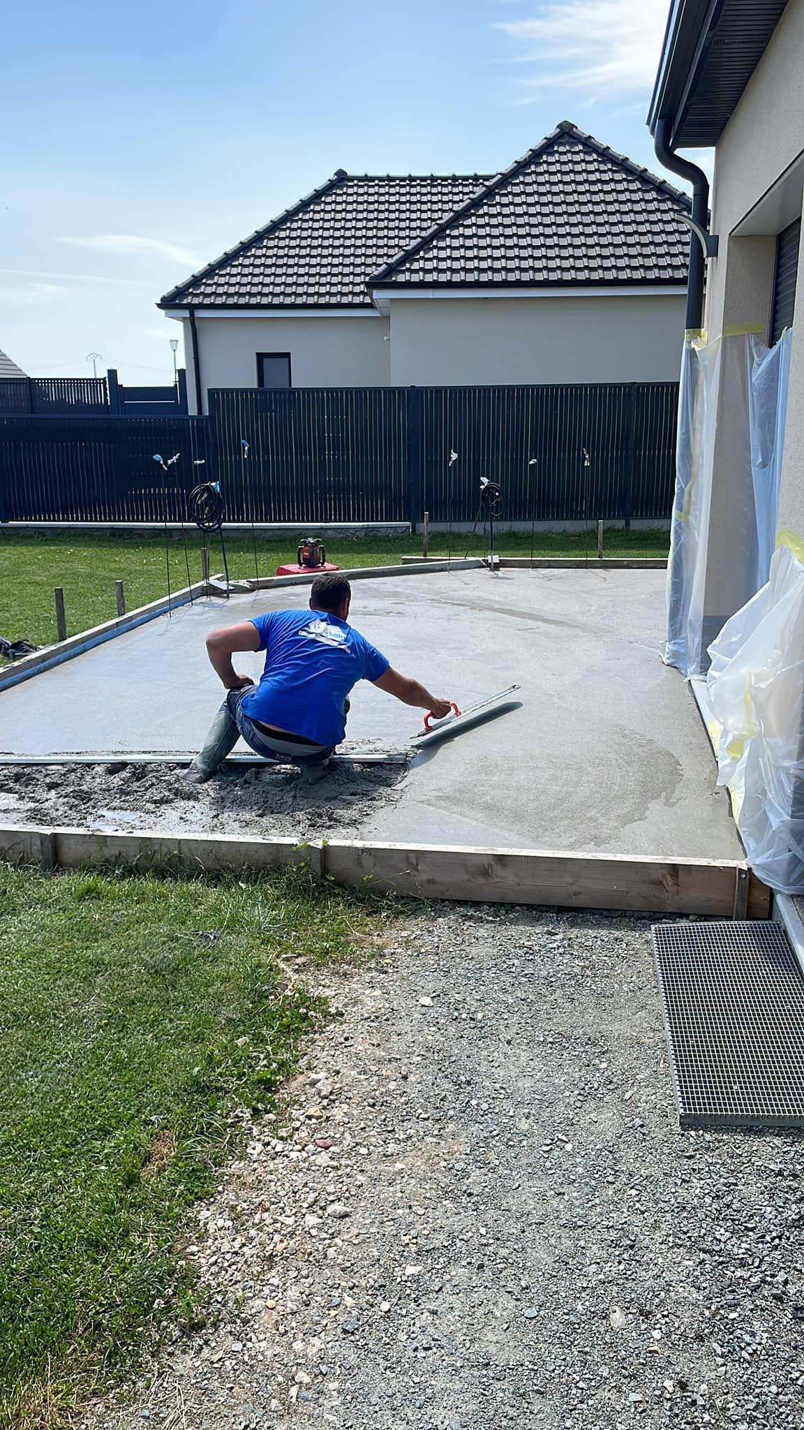 Une personne agenouillée sur une terrasse en béton, peignant un mur à côté d'une maison avec un pinceau et un bac à peinture.
