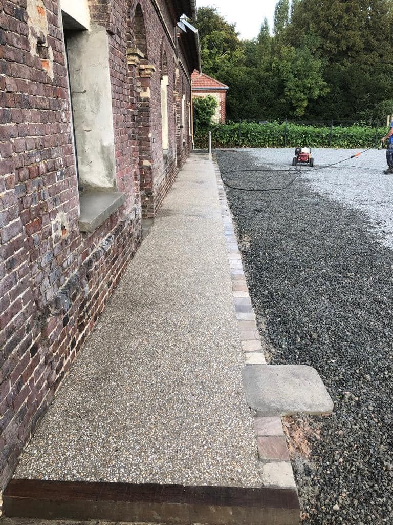 Bâtiment en briques, bordé d'un étroit chemin de gravier et d'une allée pavée, menant vers des arbres et une petite machine rouge.