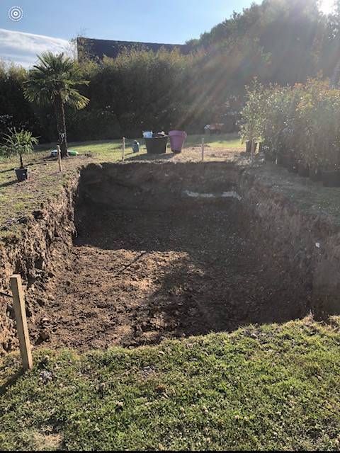 Grande excavation rectangulaire de terre dans un jardin herbeux, avec des outils et une clôture en arrière-plan.