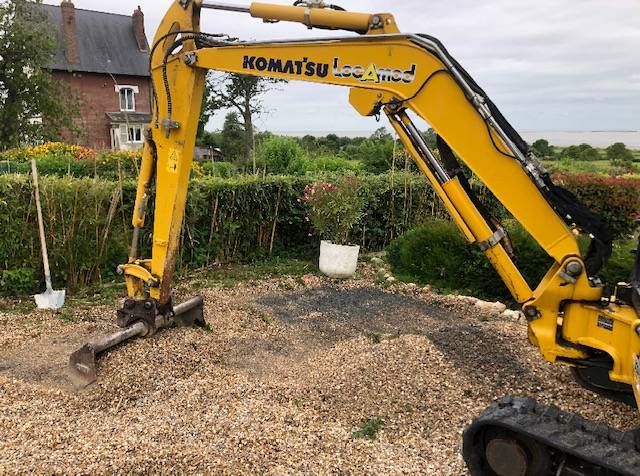 Bras d'une pelle Komatsu jaune au-dessus d'une allée de gravier, à côté d'une haie et d'une maison