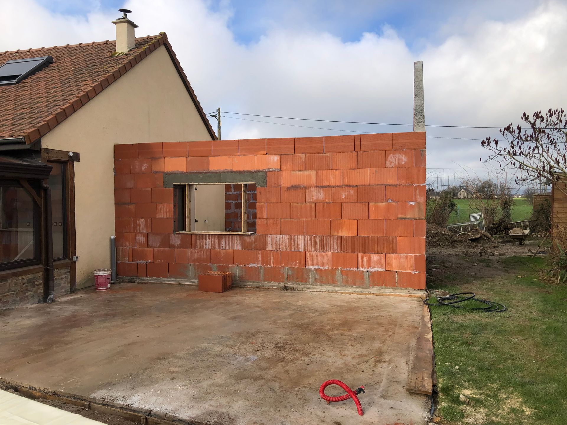 Mur en briques rouges partiellement construit à côté d'une maison, avec une terrasse en béton et une cour gazonnée sous un ciel nuageux.
