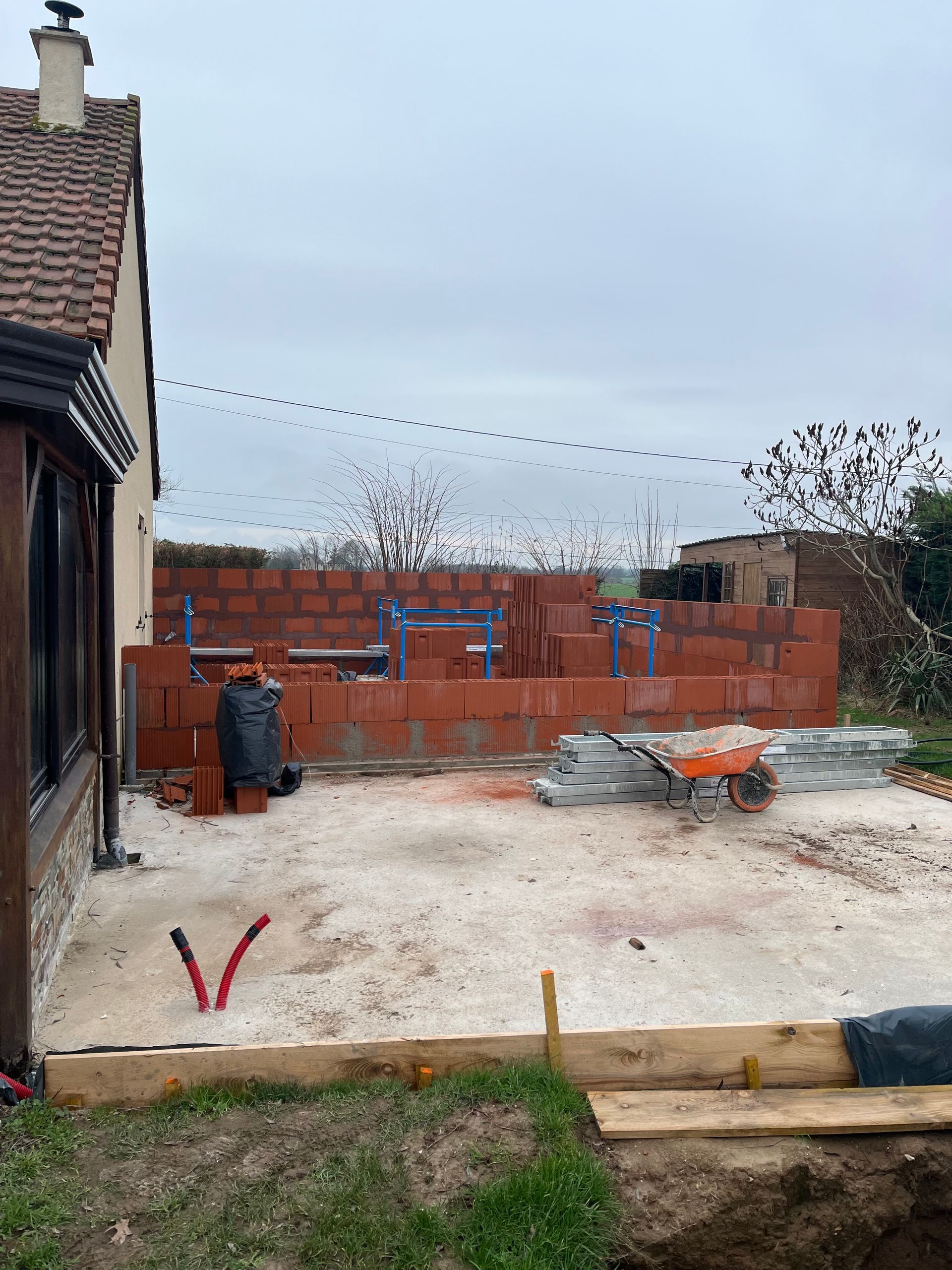 Chantier de construction dans une cour arrière avec un mur de briques inachevé, un sol en gravier et une maison sur la gauche