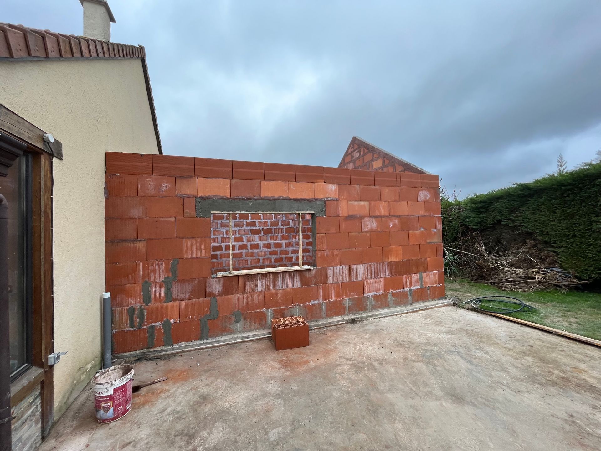 Mur en briques rouges partiellement construit à côté d'une maison et d'une terrasse en béton sous un ciel nuageux