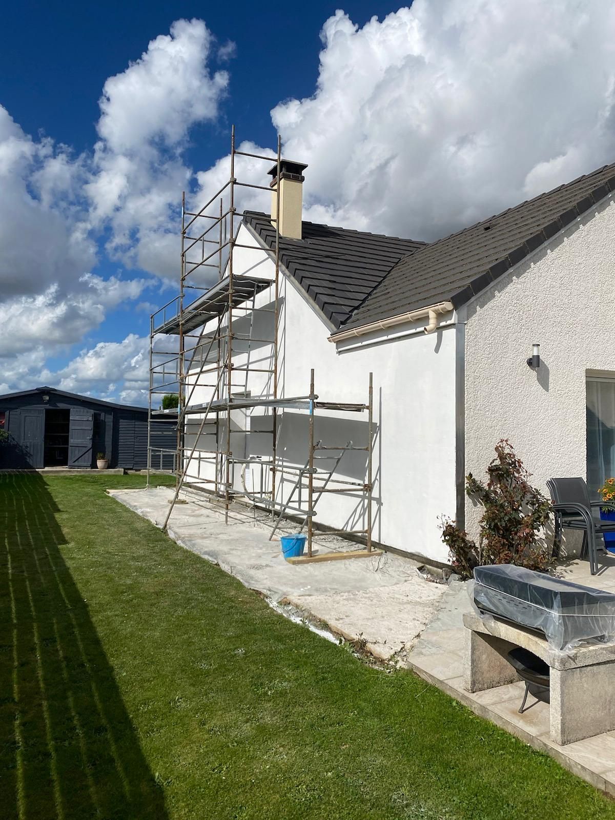 Échafaudages le long de la façade d'une maison blanche, à côté d'une pelouse verte sous un ciel partiellement nuageux