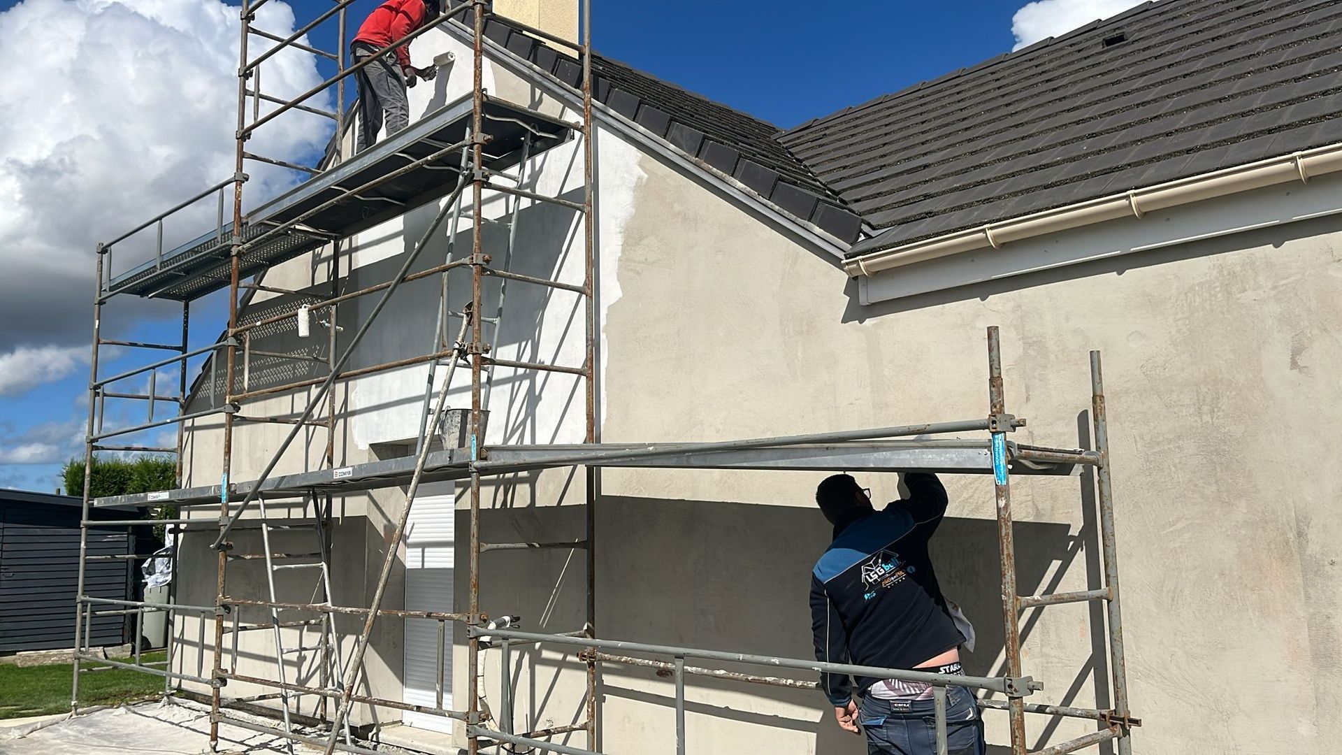 Des ouvriers, perchés sur des échafaudages, peignent la façade d'une maison blanche sous un ciel bleu.
