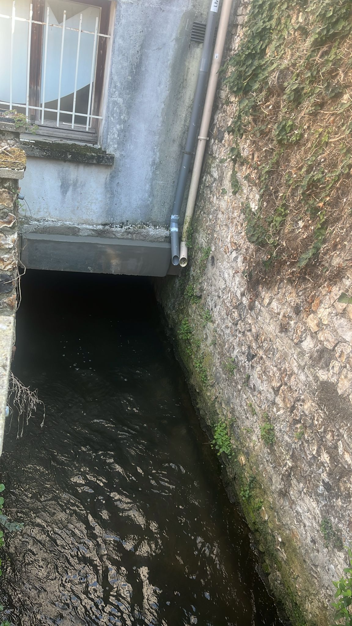 Canal étroit longeant un mur de pierre grise, aux eaux sombres et surmonté d'une petite structure métallique.