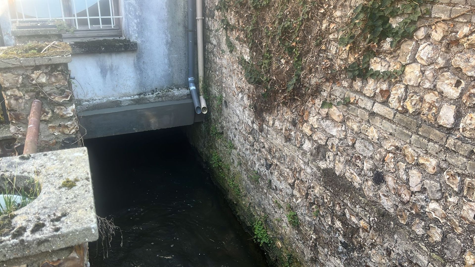 Un étroit canal longeant un mur de pierre et un pont en béton dans un cadre champêtre.