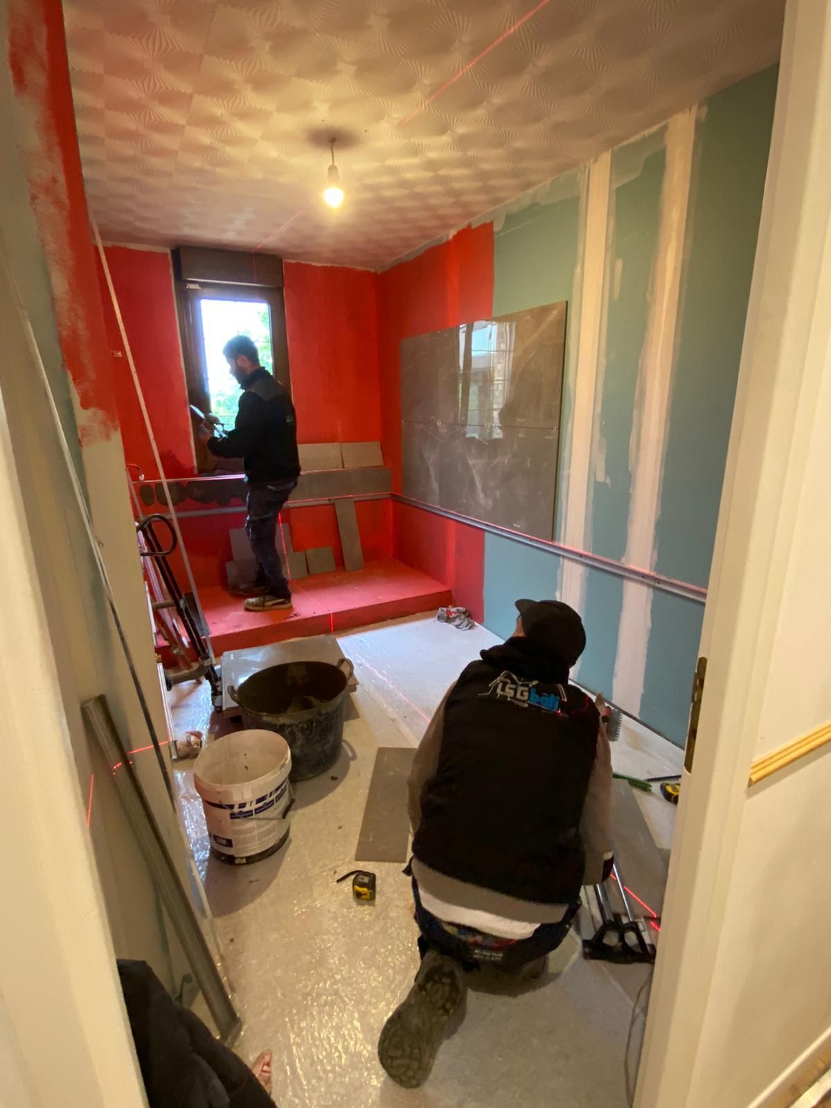Deux ouvriers peignent un couloir étroit, aux murs rouges et blancs, avec des outils éparpillés sur le sol.
