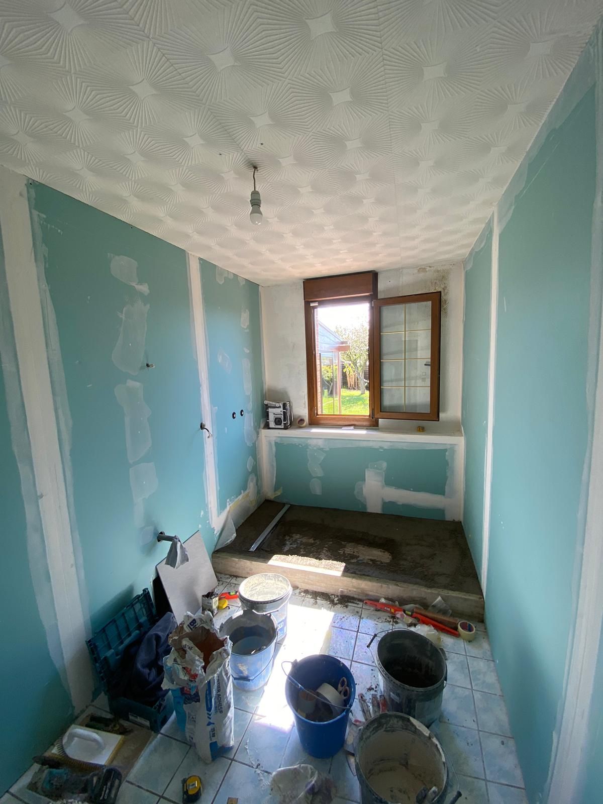 Pièce étroite en rénovation, aux murs bleus, avec des seaux de peinture au sol et une fenêtre au fond.