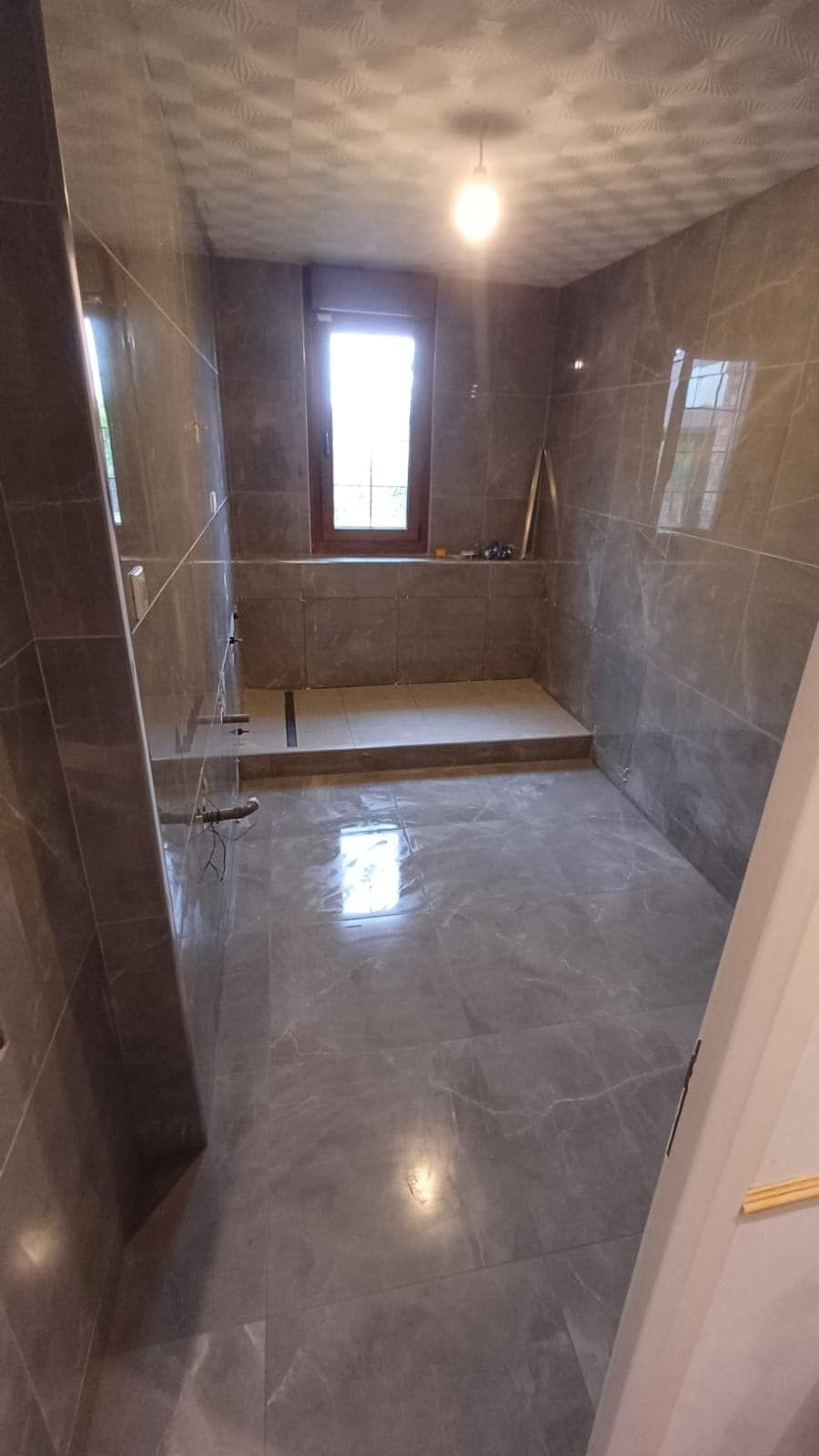 Salle de douche carrelée vide, avec murs gris brillants, banc et petite fenêtre.