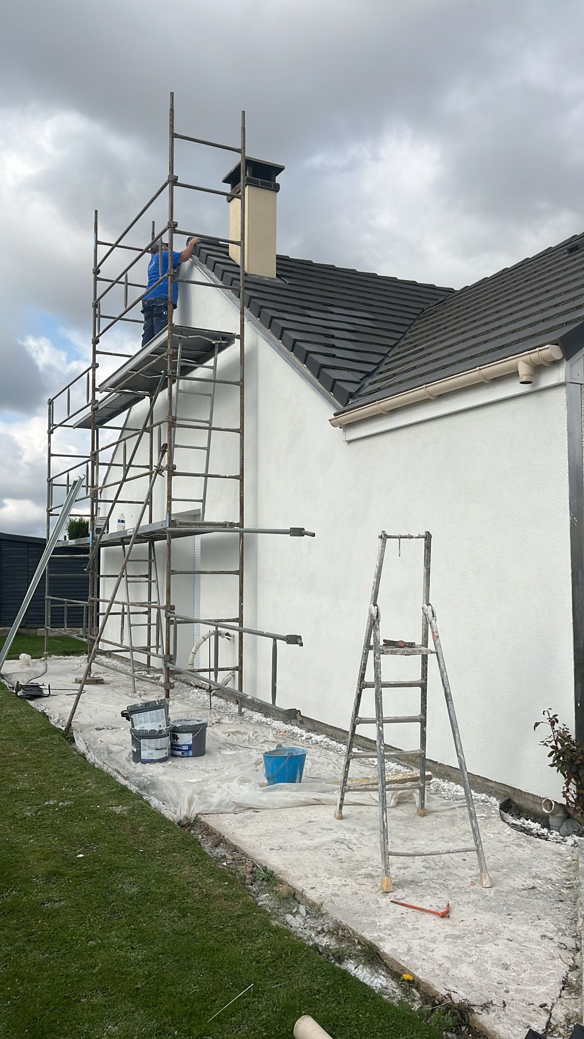 Échafaudages à côté d'une maison blanche, un ouvrier peint la partie supérieure du mur sous un ciel nuageux.