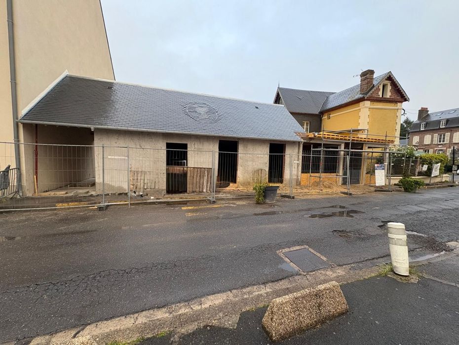 Bâtiment en béton gris inachevé, avec une grande porte, des tas de terre devant et une petite excavatrice sur la droite.