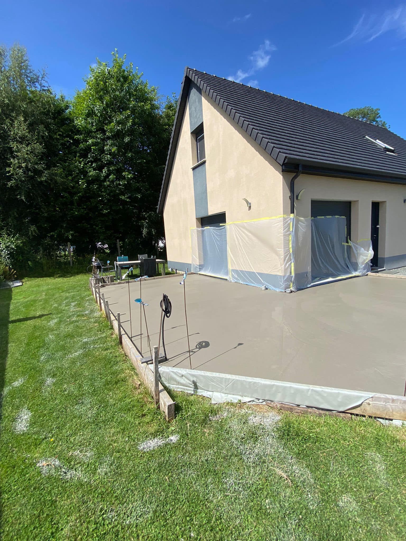 Maison moderne aux murs beiges, au toit foncé et à la cour clôturée bordée d'herbe verte et d'arbres.