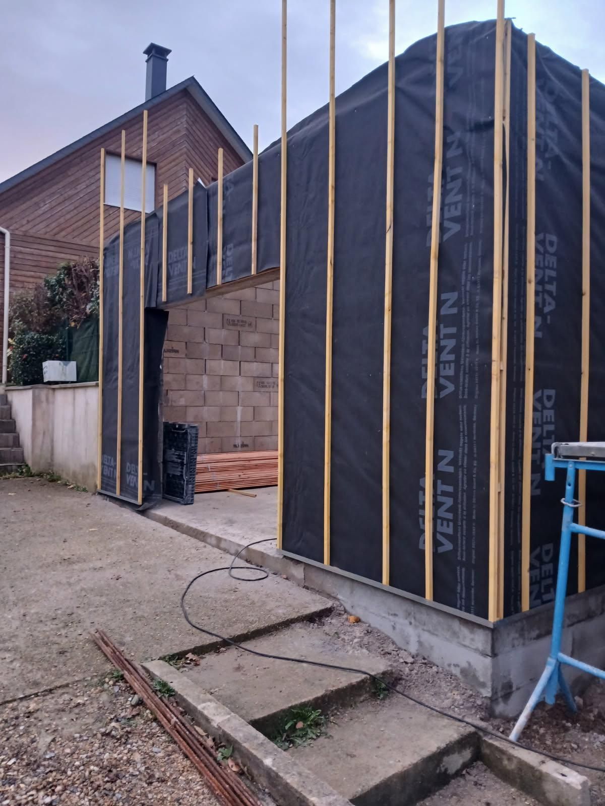 Extérieur d'une maison partiellement construite, avec revêtement noir, charpente apparente en bois et petite marche d'entrée en béton.