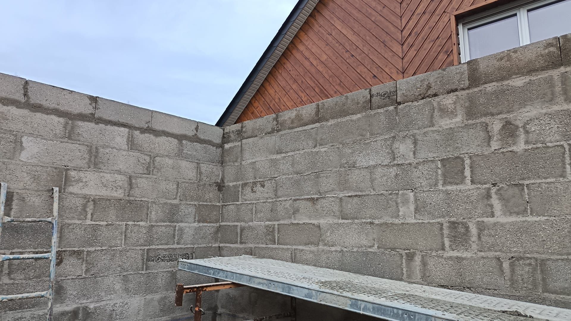 Coin d'un mur en parpaings avec un banc ou une table en métal sous le pignon et la fenêtre d'une maison en briques