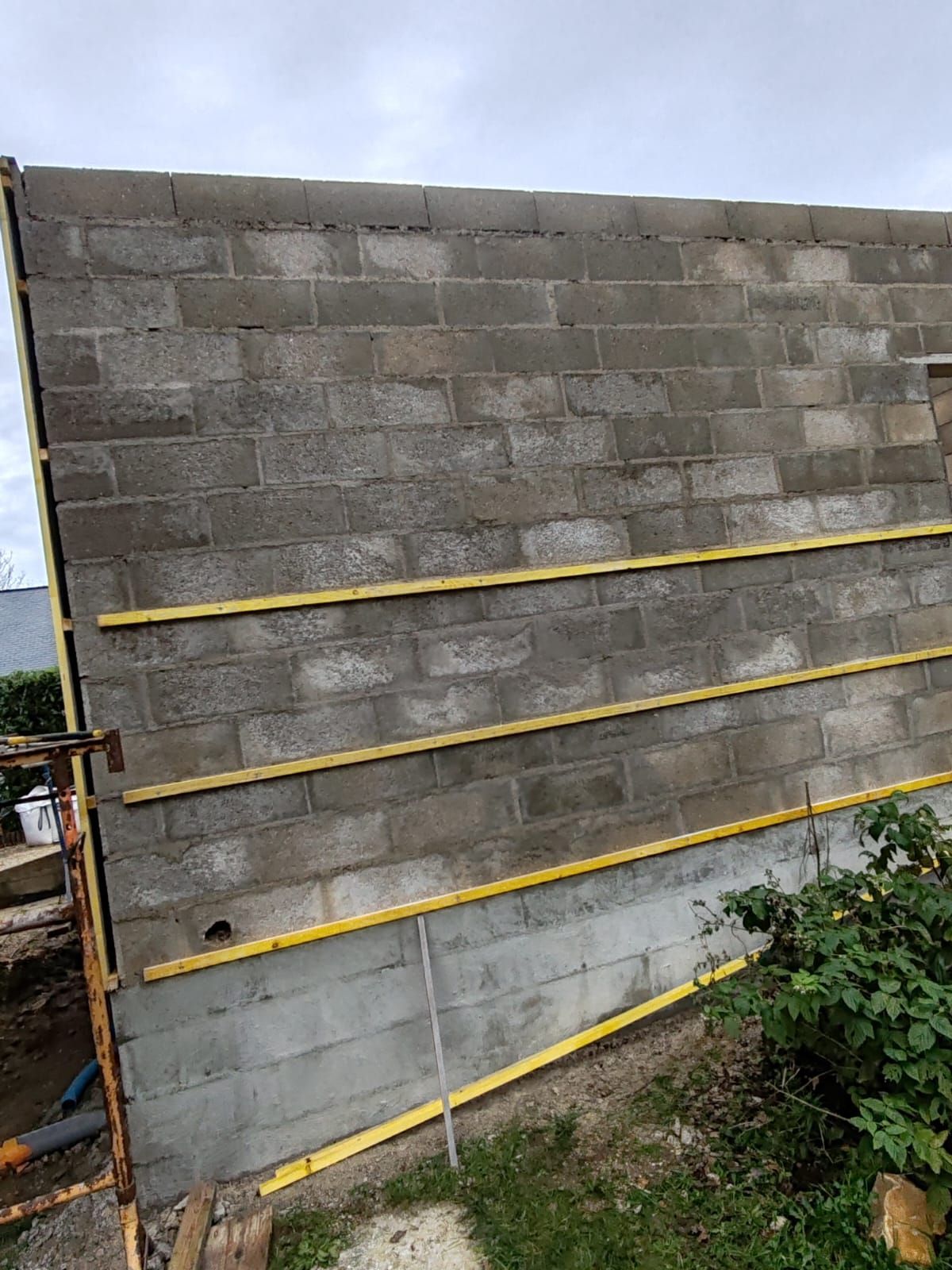 Mur en parpaings de béton avec trois entretoises horizontales jaunes dans une cour extérieure