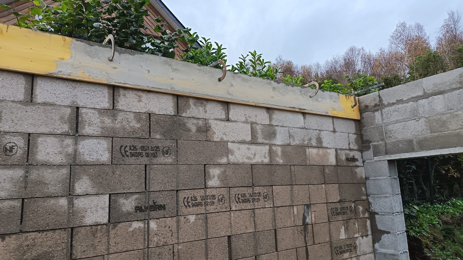 Mur en parpaings gris partiellement construit, avec une assise supérieure jaune et une ouverture pour la fenêtre.