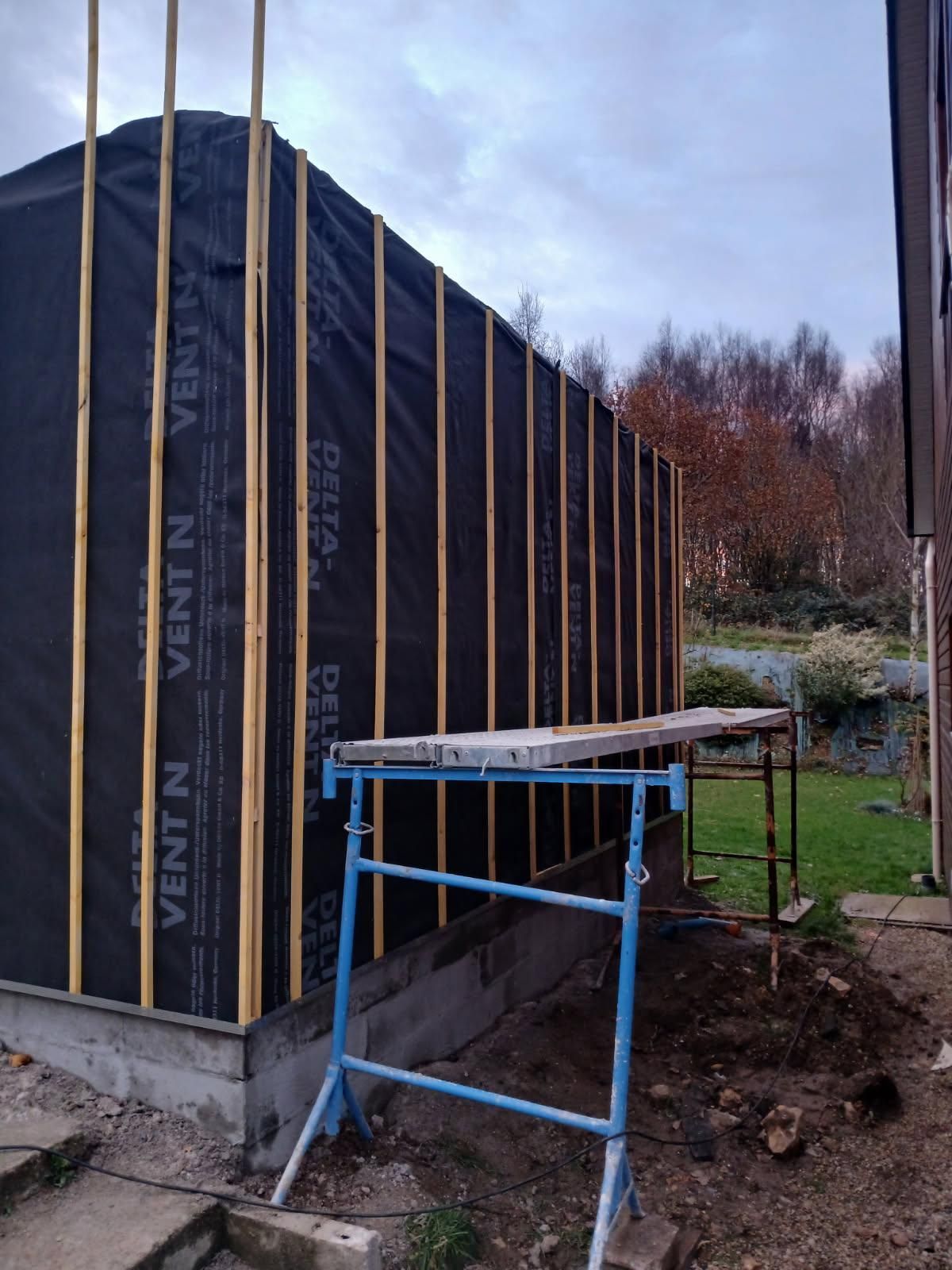 Structure en bois partiellement construite, recouverte d'un revêtement noir, avec des échafaudages bleus dans une cour boueuse.