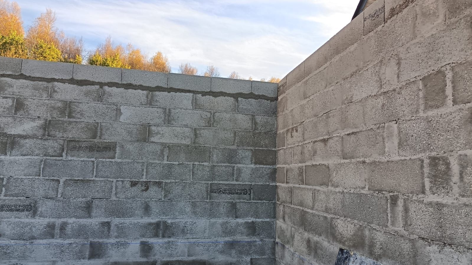 Des murs en parpaings de béton forment un coin à l'extérieur sous un ciel nuageux, avec des arbres d'automne visibles au-dessus.