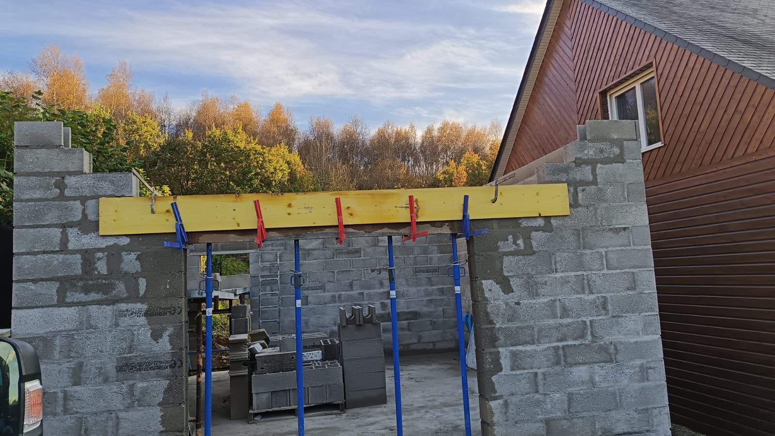 Chantier de construction avec murs en parpaings, poutre jaune et supports bleus à côté d'un bâtiment en briques