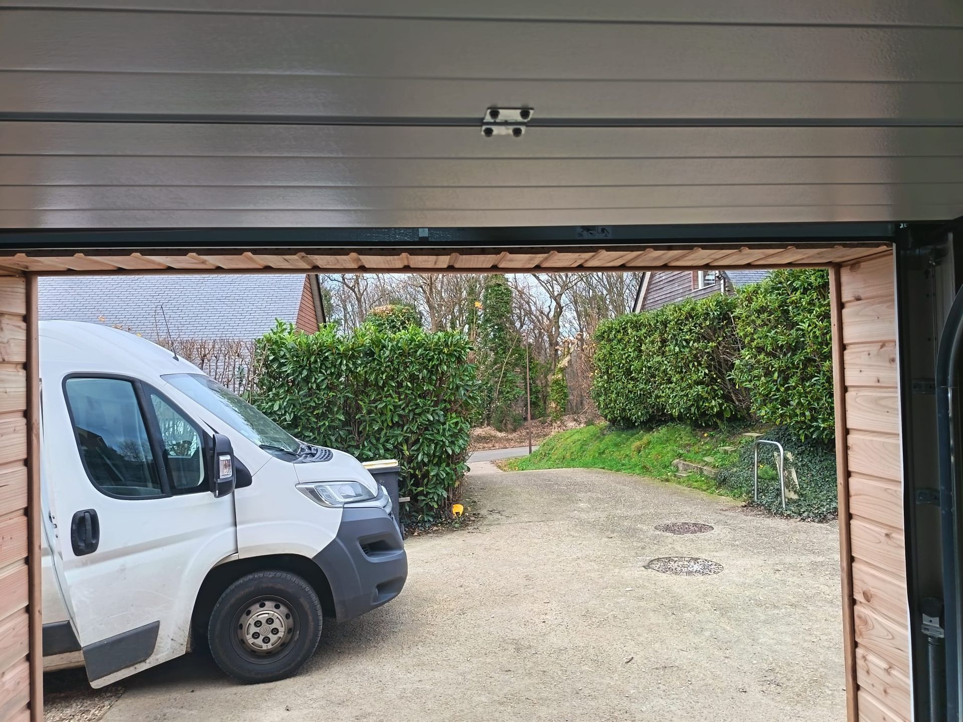 Vue depuis un garage montrant une camionnette blanche garée dans une allée, avec des buissons et des arbres au-delà.