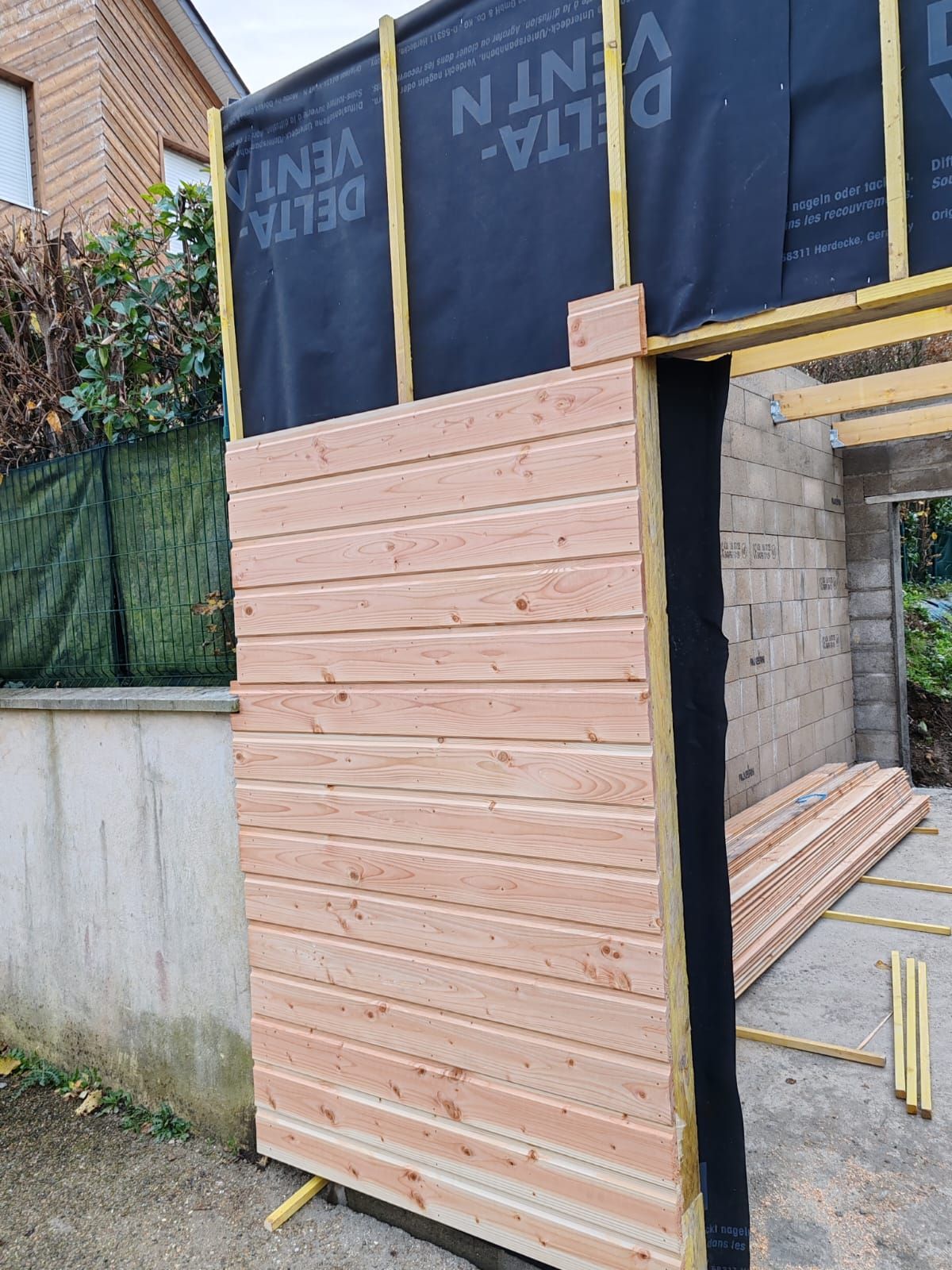 Mur en bois partiellement construit, avec des planches roses et un pare-vapeur noir, sur un chantier.