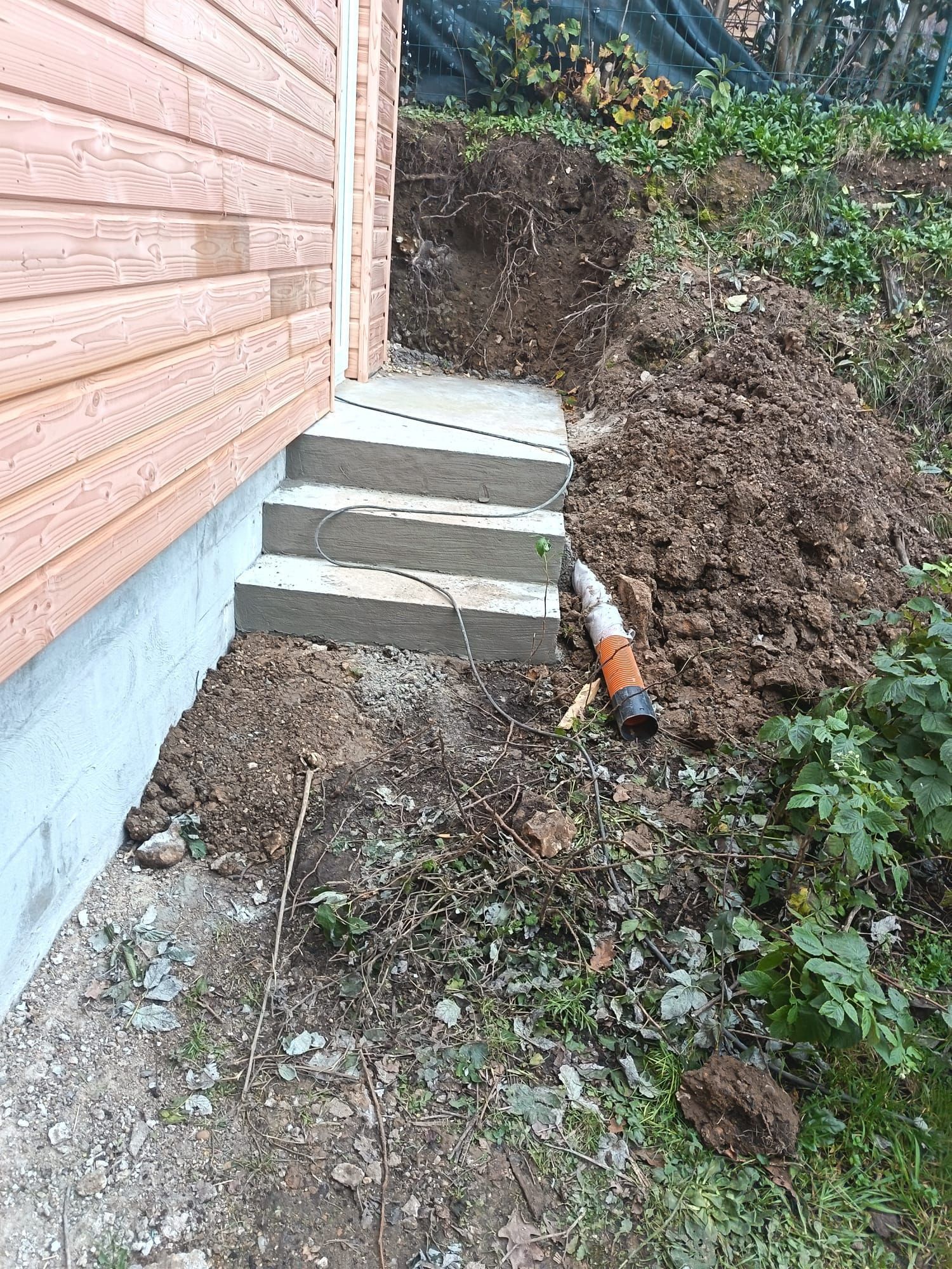 Escaliers en béton partiellement construits à côté d'une maison, avec de la terre et des débris amoncelés sur le côté.