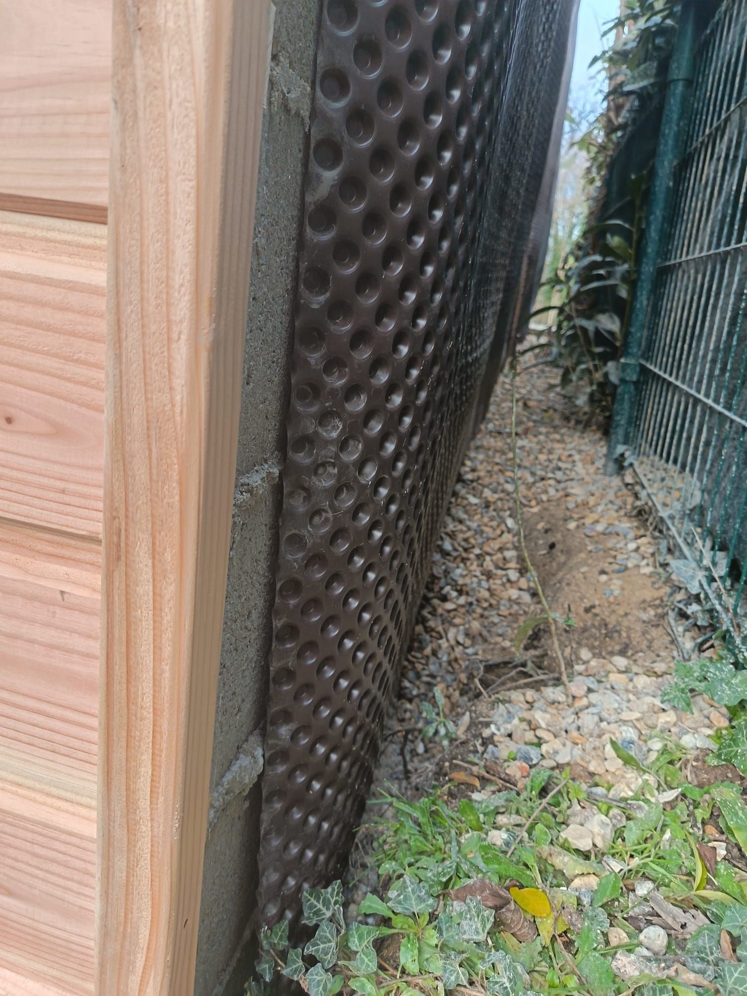 Étroit passage extérieur entre un mur en bois et une clôture noire perforée, avec du gravier et des feuilles au sol.