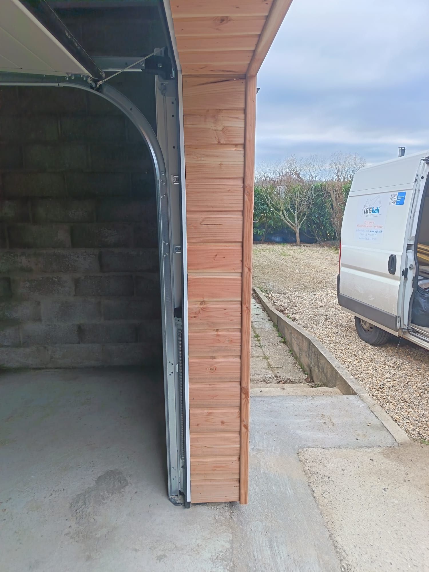 Porte de garage ouverte à côté d'un bardage en bois, avec une camionnette blanche garée sur une allée de gravier.