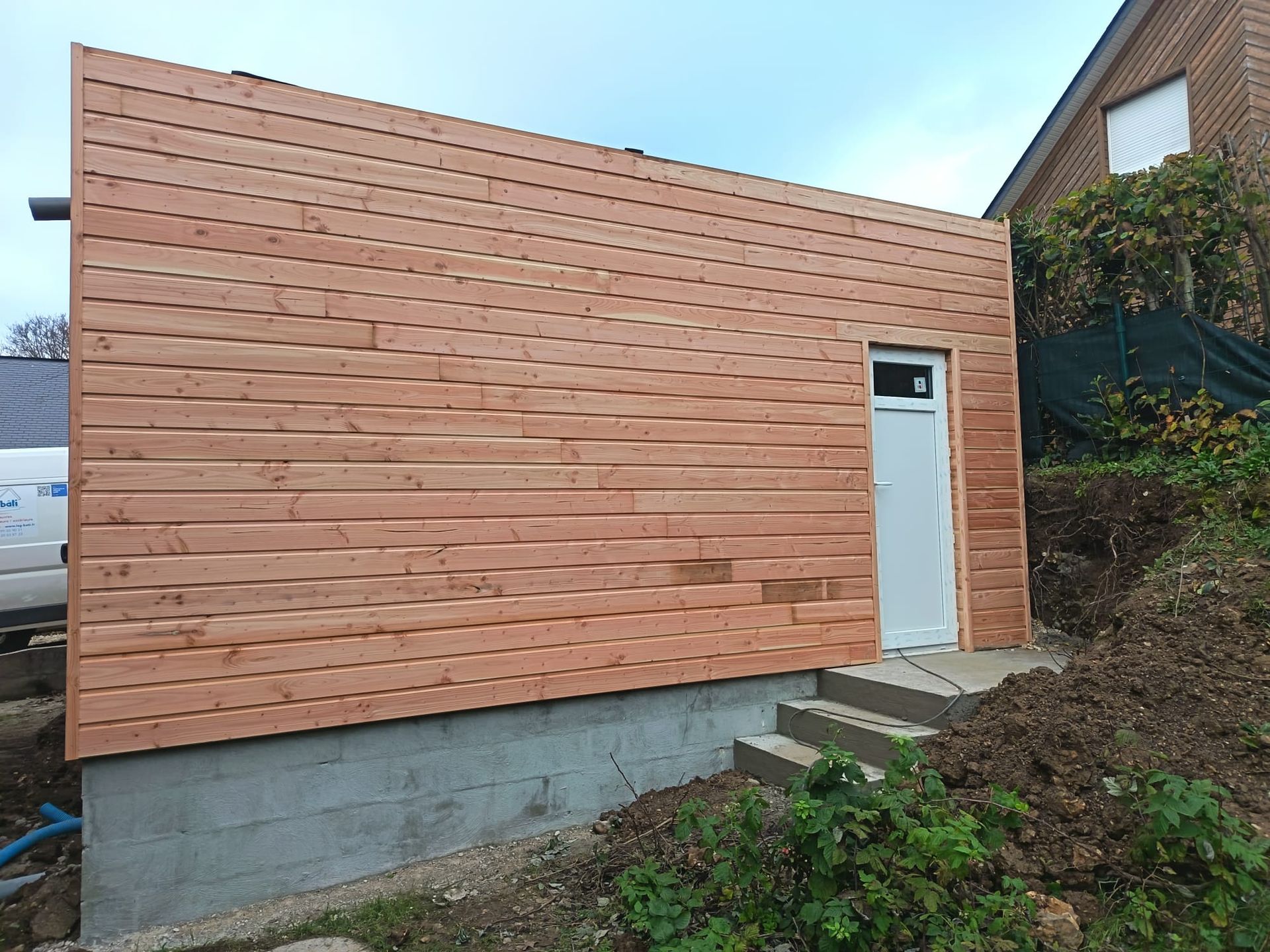 Façade d'un bâtiment revêtu de bois avec une porte blanche sur des fondations en béton à flanc de colline.