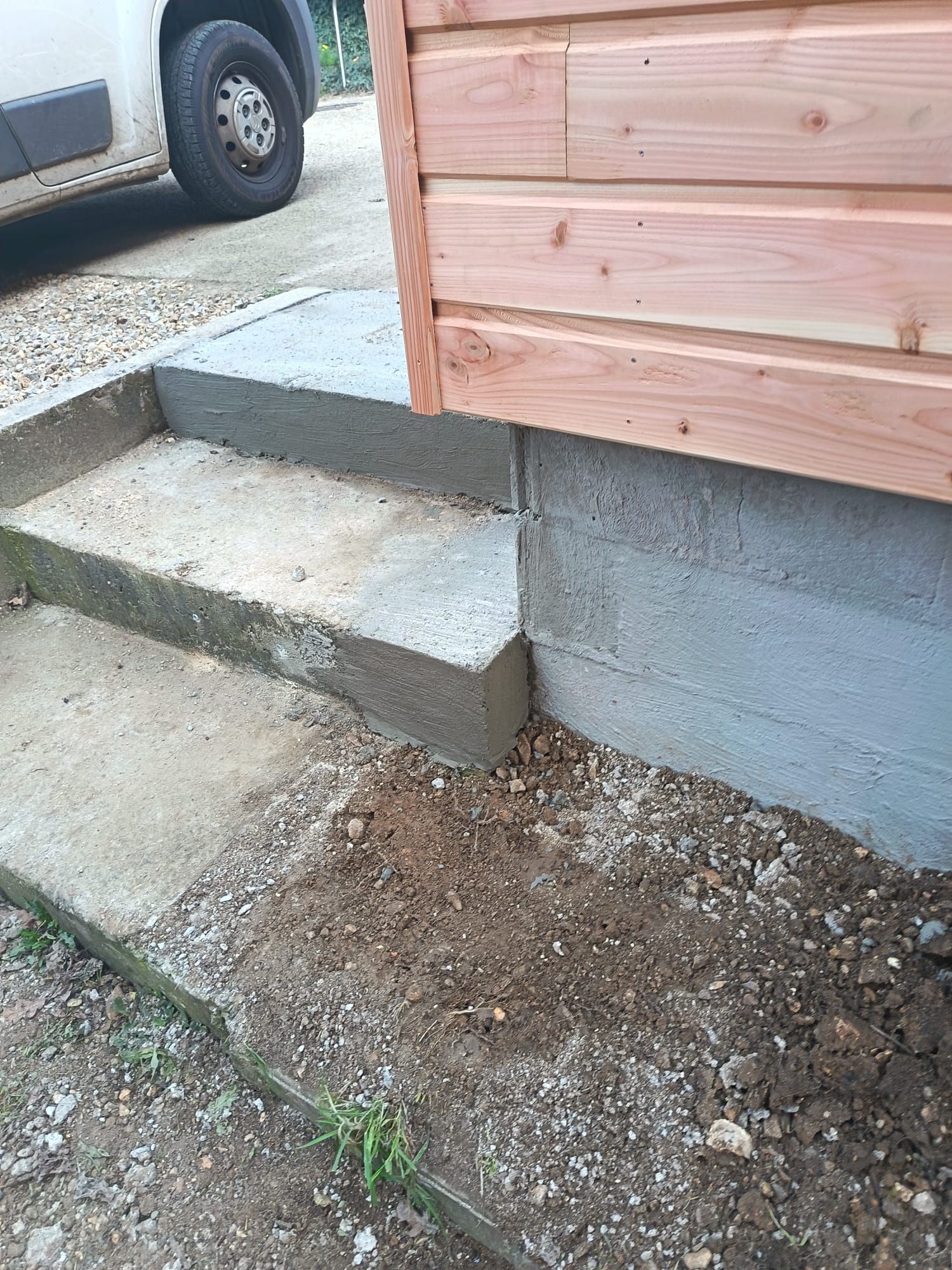 Des marches en béton mènent à un abri en bois situé à côté d'un terrain vague, avec un pneu de véhicule visible en arrière-plan.