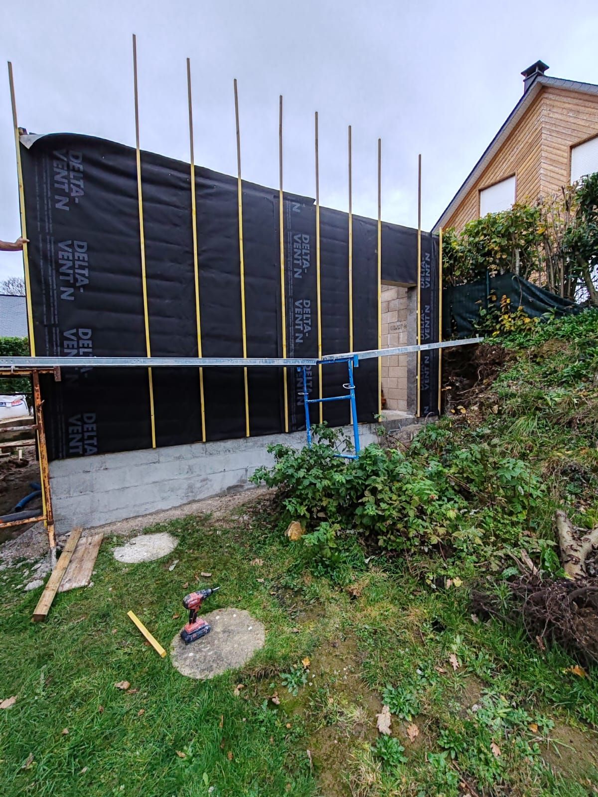 Façade d'une maison en construction, recouverte d'un pare-vapeur noir, avec des montants en bois et un échafaudage bleu devant une cour en pente.