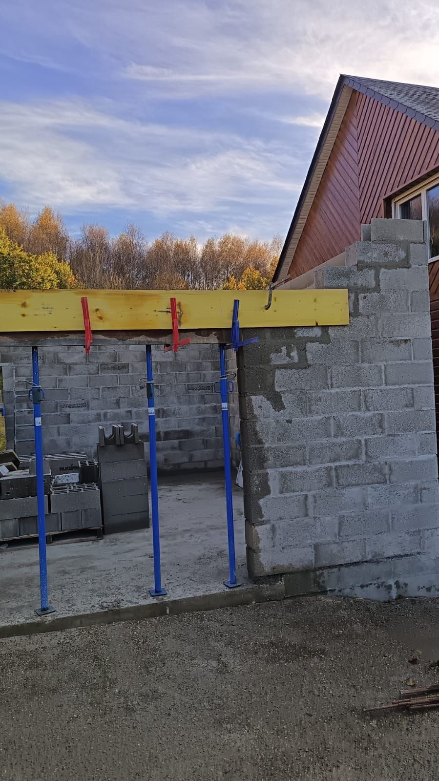 Mur en parpaings partiellement construit, avec poutre jaune et poteaux de soutien bleus, sous un ciel nuageux.