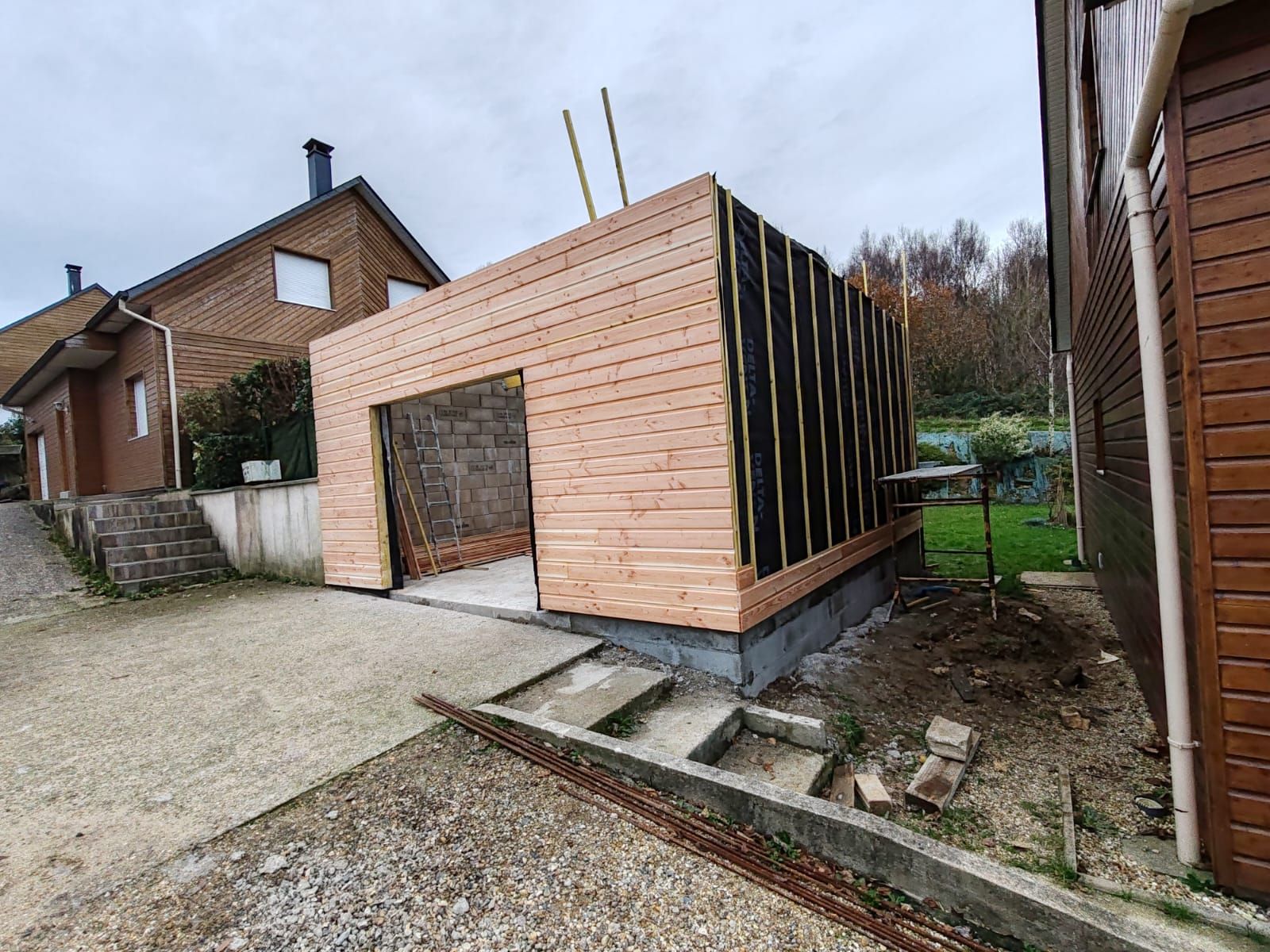 Abri de jardin en bois partiellement construit, avec façade ouverte, situé dans une cour gravillonnée à côté de maisons.