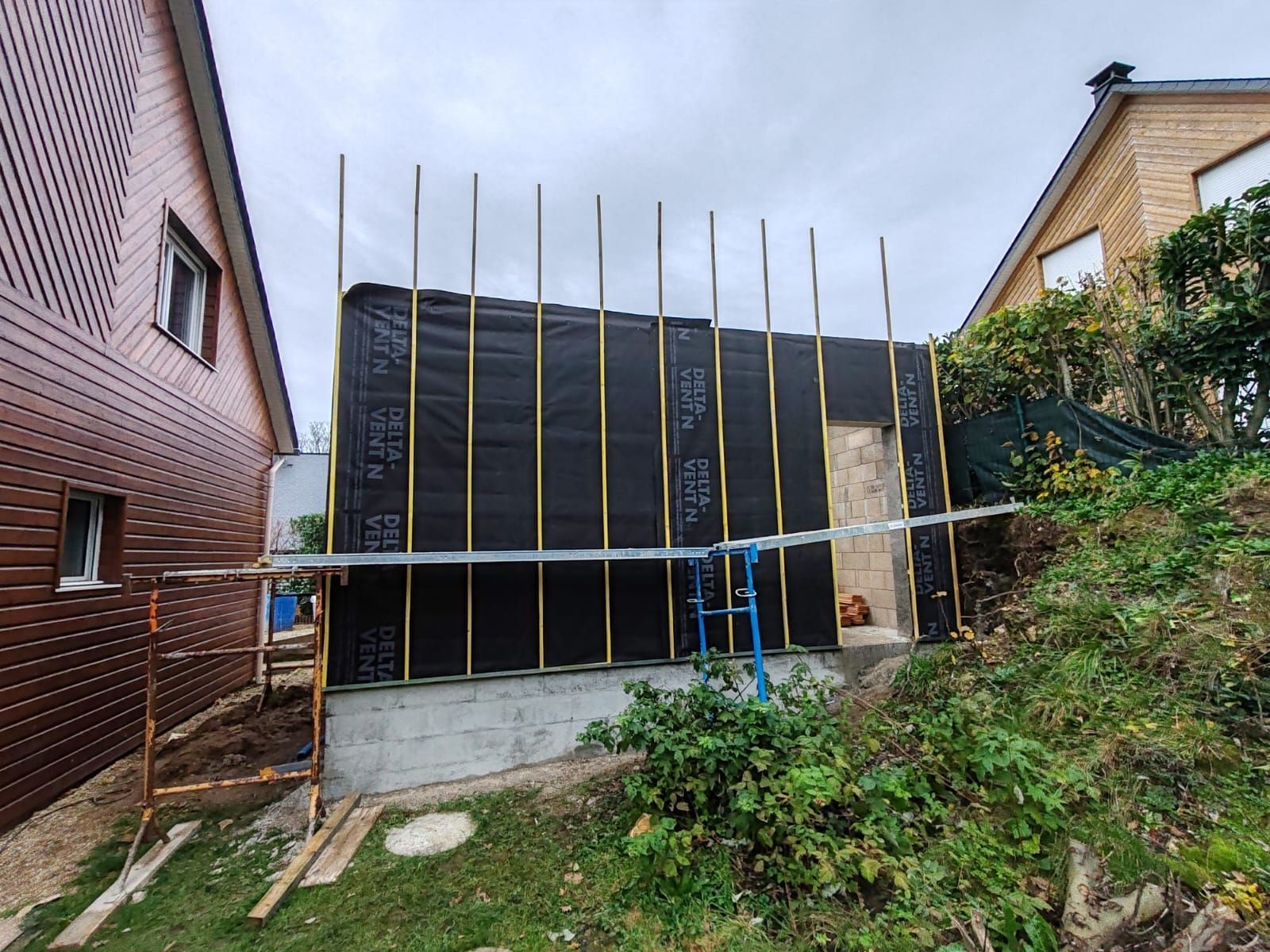 Extérieur d'une maison partiellement construite avec un revêtement noir, des poteaux verticaux et un échafaudage entre deux maisons