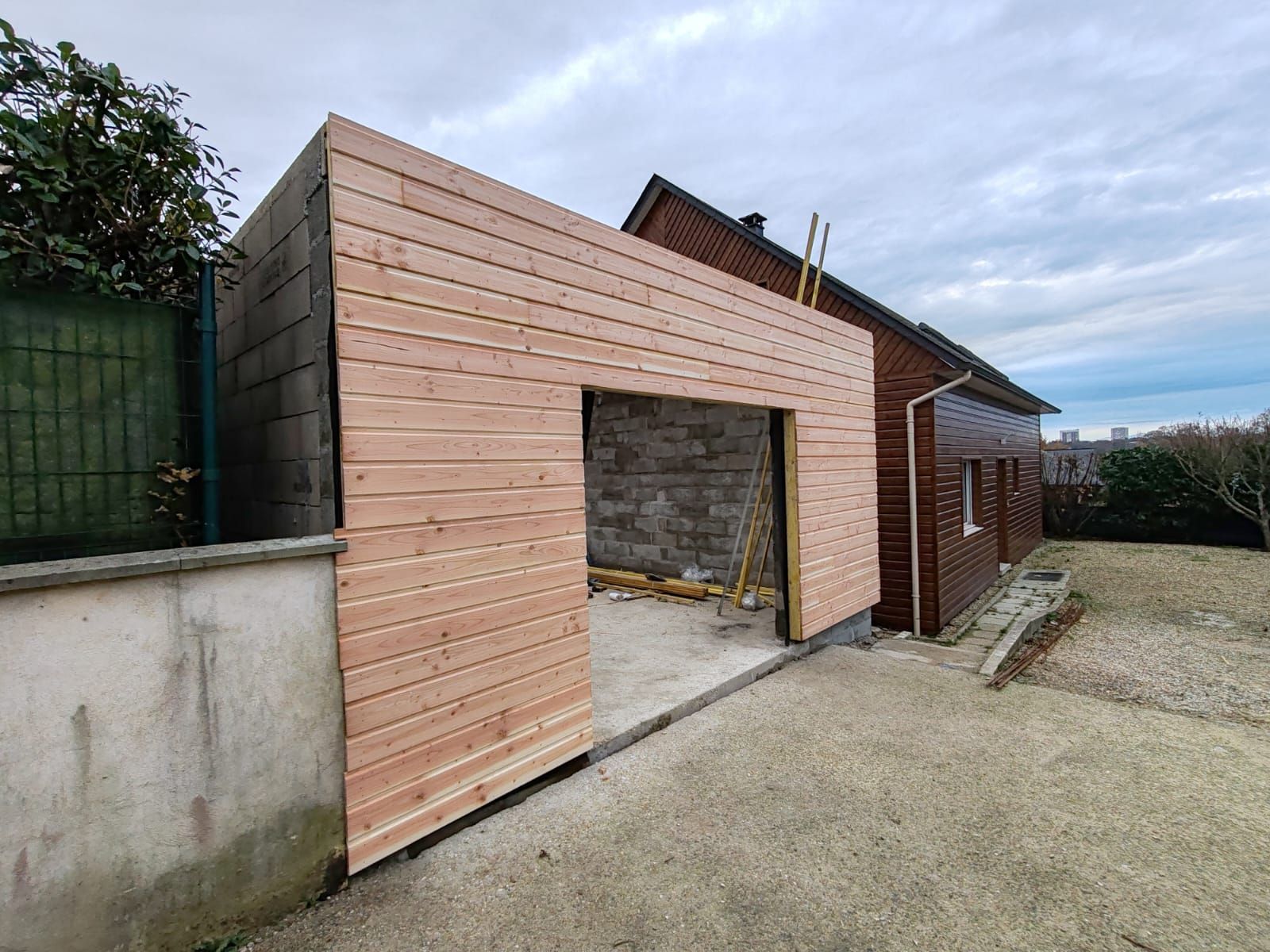Extension de garage moderne en bois avec une grande ouverture à côté d'une maison, visible depuis une allée gravillonnée.
