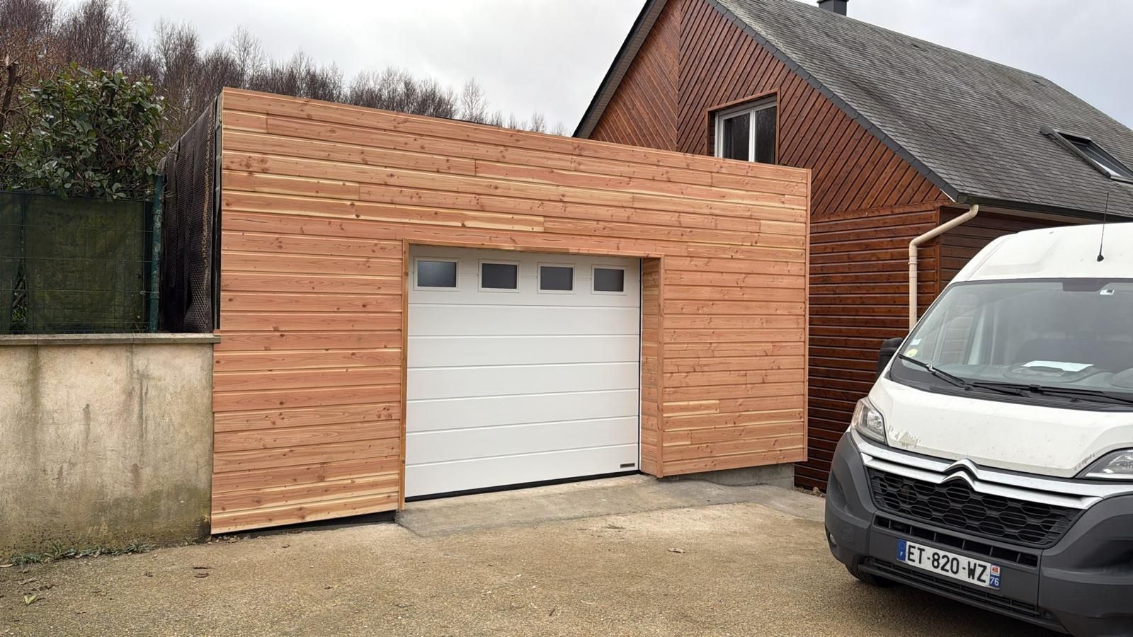Garage indépendant revêtu de bois avec porte blanche, à côté d'une camionnette blanche garée dans une allée résidentielle.