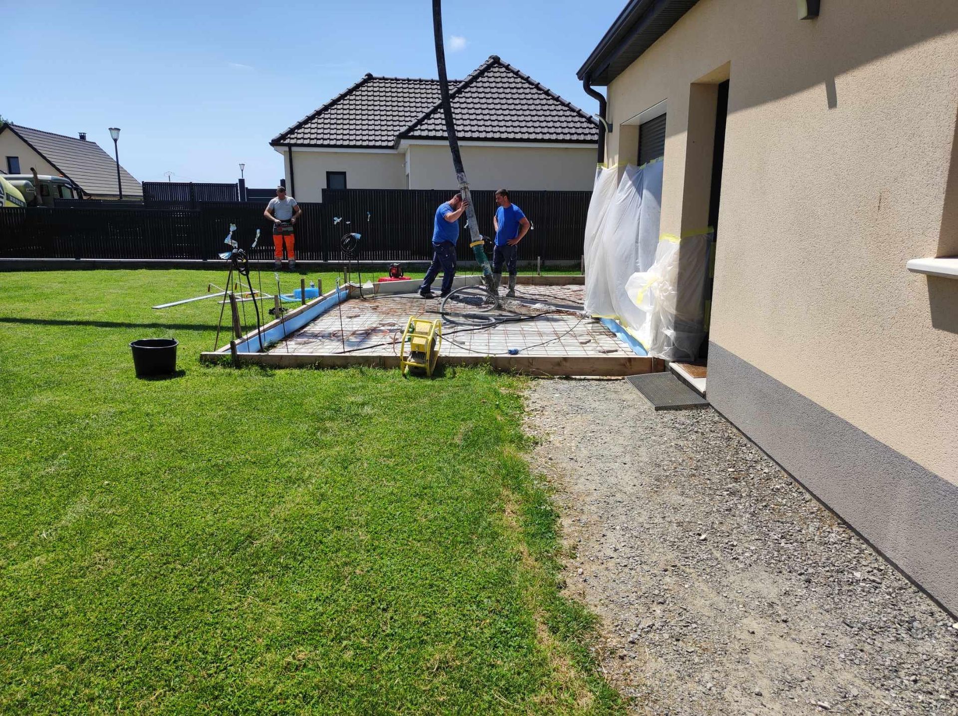 Des ouvriers installent une terrasse à côté d'une maison, avec leurs outils et matériaux, sur une pelouse ensoleillée.