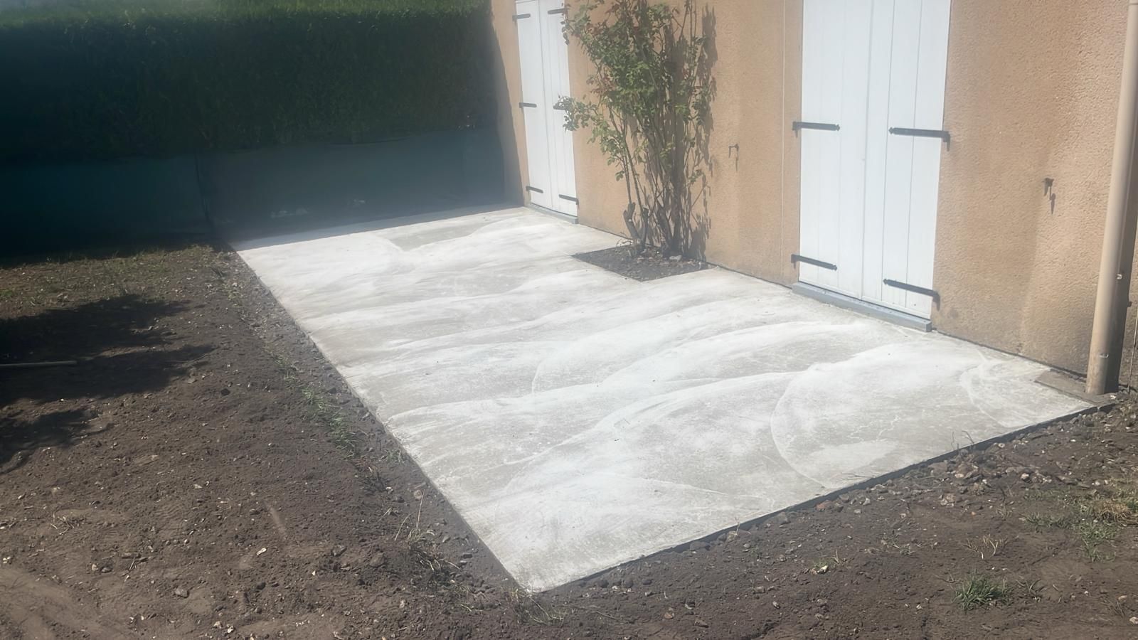Terrasse en béton jouxtant un bâtiment beige aux portes blanches, bordée de terre sombre et d'un palmier.