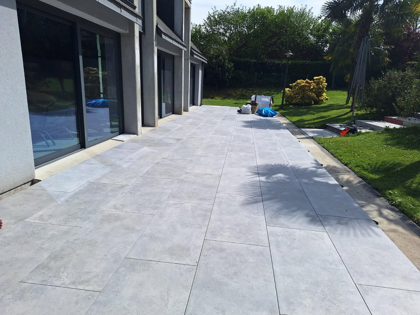 Terrasse en béton jouxtant une maison moderne, avec outils et matériaux près d'un jardin gazonné.