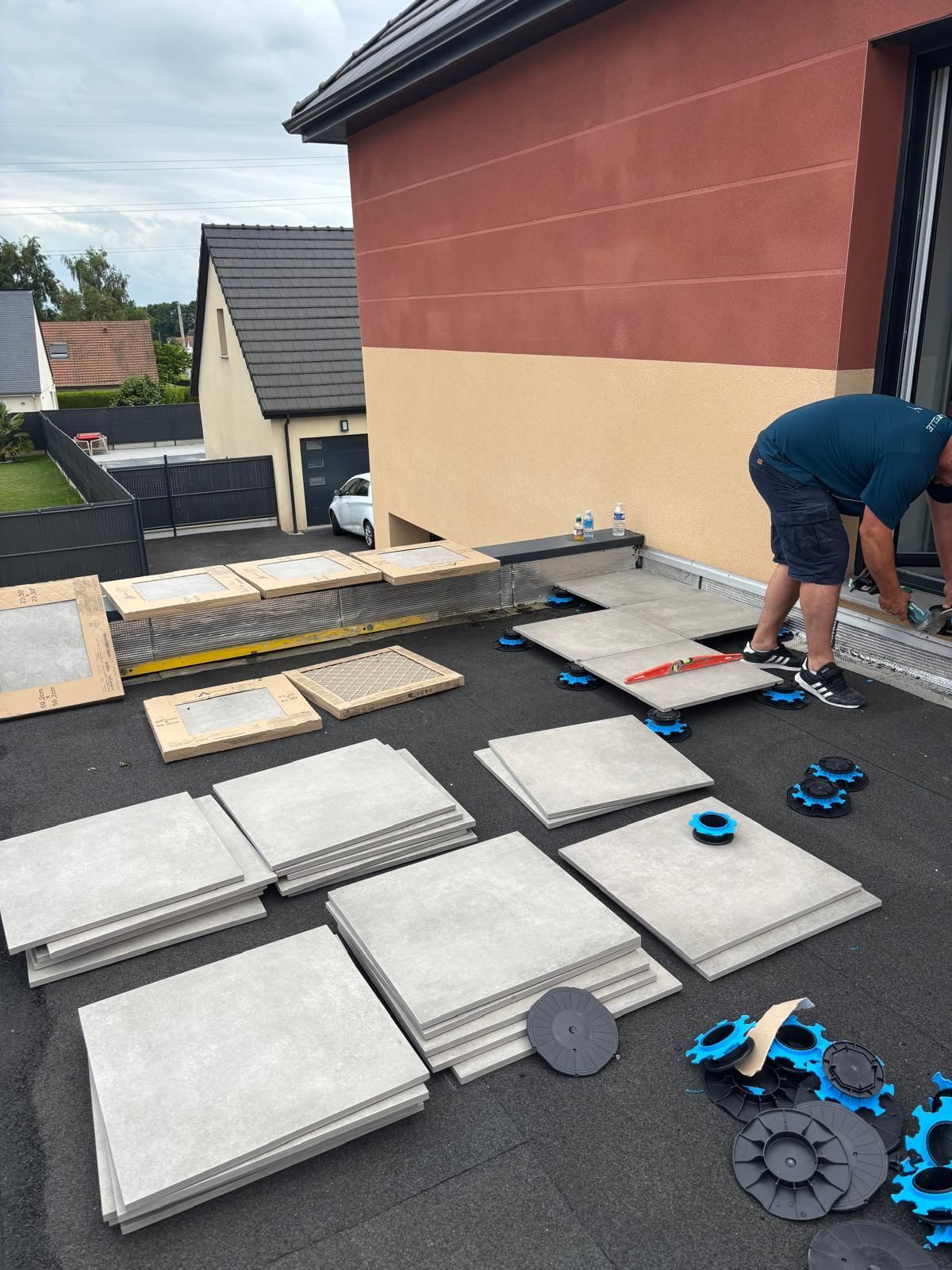 Un ouvrier installe des dalles de patio gris clair sur une terrasse de toit, à côté d'une maison.