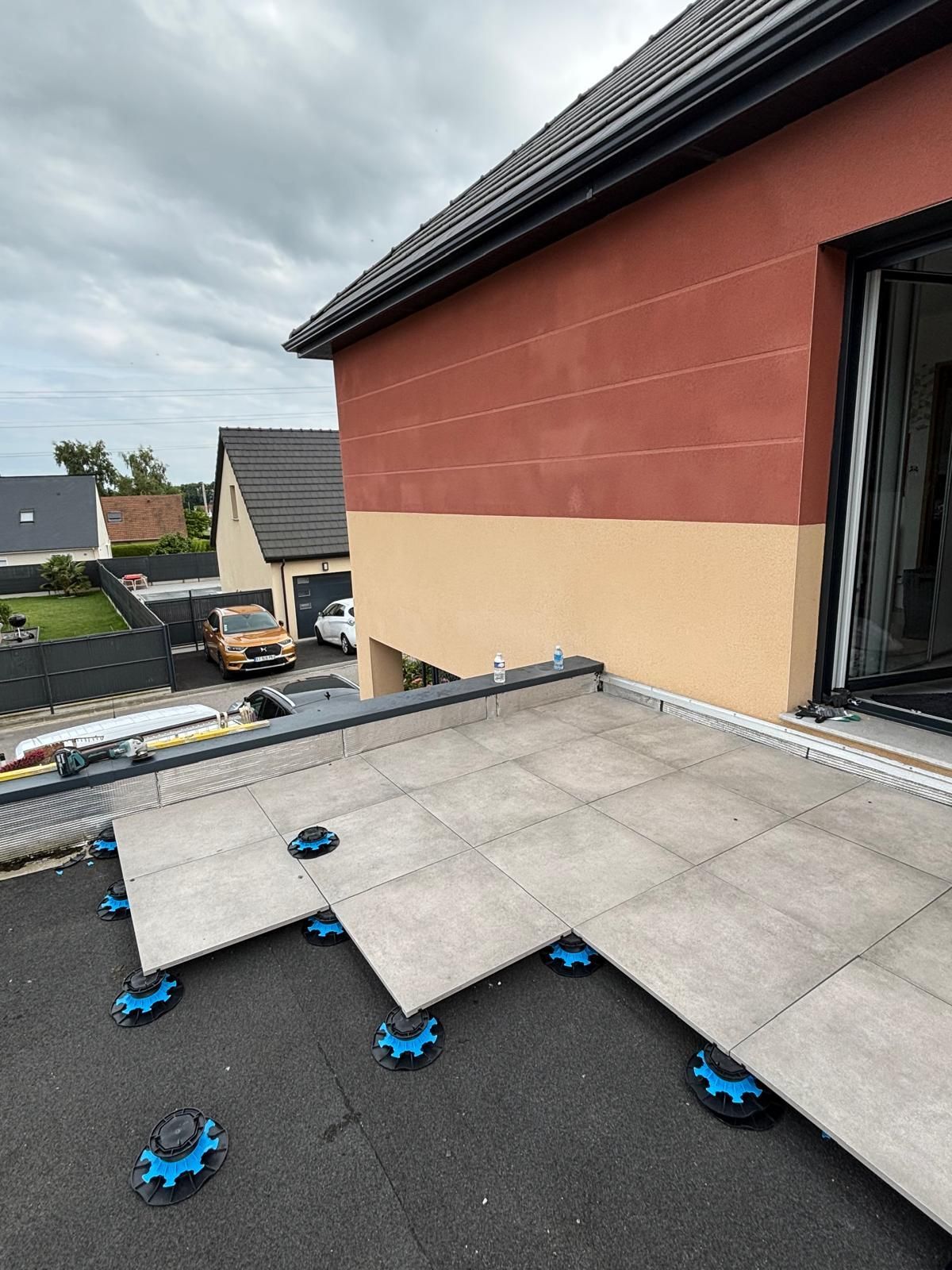 Terrasse sur toit plat en construction avec des tuiles grises et des cales de nivellement bleues à côté d'une maison rouge et beige