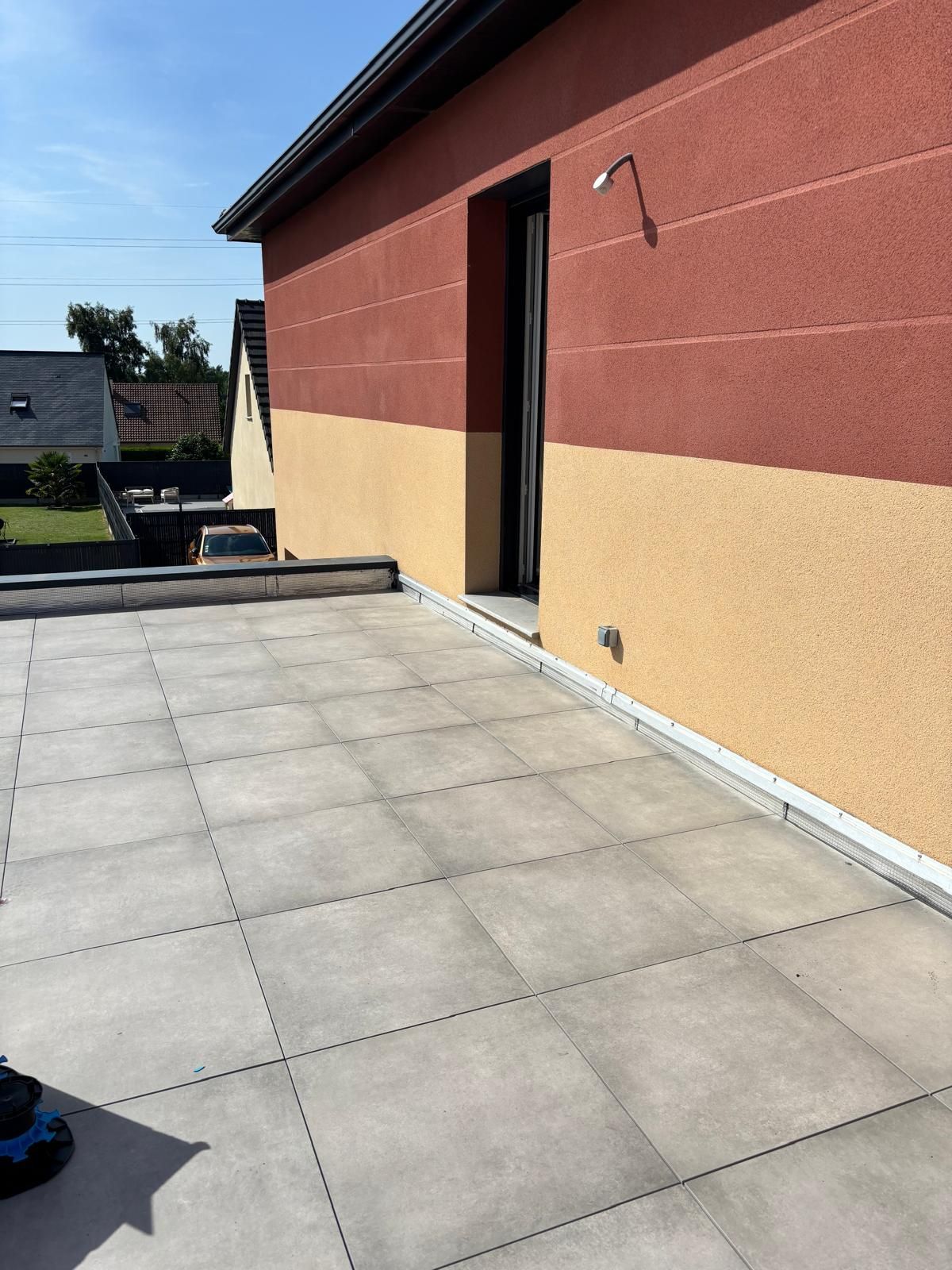 Terrasse extérieure carrelée de gris, jouxtant un bâtiment rouge et beige et une porte ouverte.