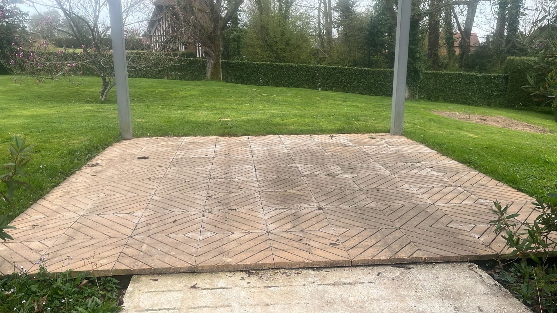 Dalle pavée de briques avec poteaux métalliques dans un parc gazonné, bordé d'arbres et d'un trottoir.