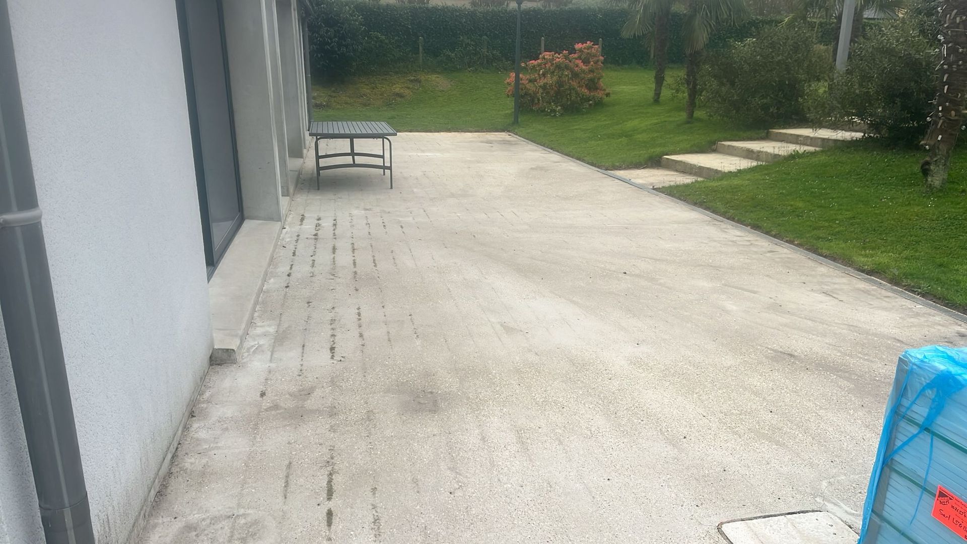 Terrasse en béton jouxtant une maison grise, avec un banc et une pelouse sous un ciel nuageux.