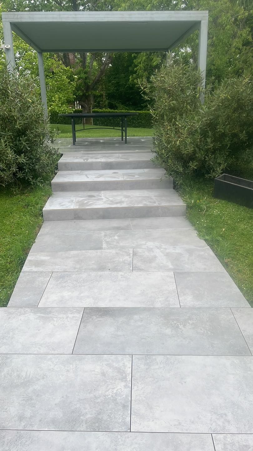 Un chemin de pierre mène à une pergola ombragée avec des bancs, bordée d'arbustes et de pelouse.